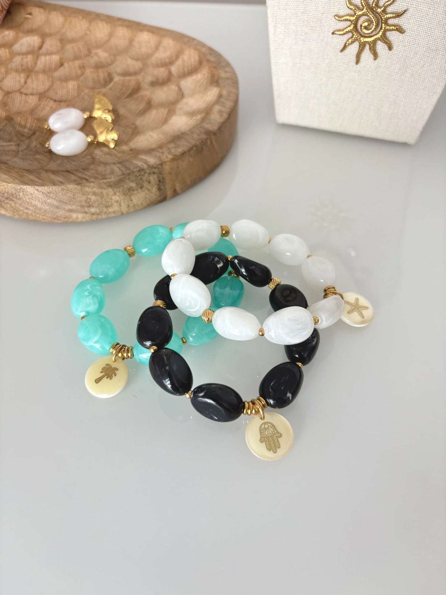 Trois bracelets superposés à élastique composés de grandes perles colorées turquoise, blanches et noires. Bijoux ornés d'un sequin en nacre blanche et ornés d'un motif doré palmier, étoile de mer et main de fatma.