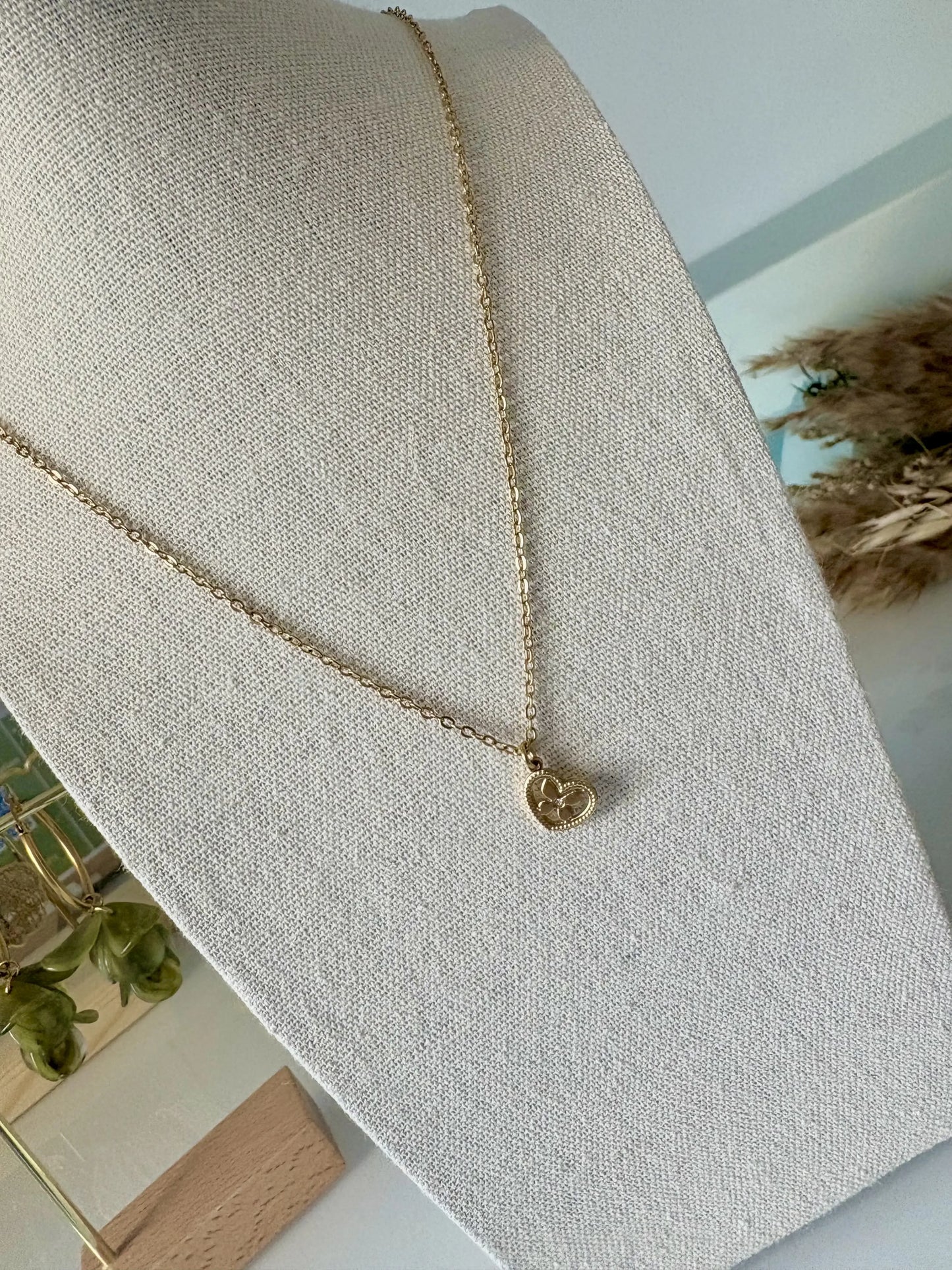 Collier à chaîne fine orné d'un pendentif coeur imbriqué d'un papillon décoré d'un strass en son centre. Bijou en acier inoxydable doré. Bijou posé sur un buste en coton beige.
