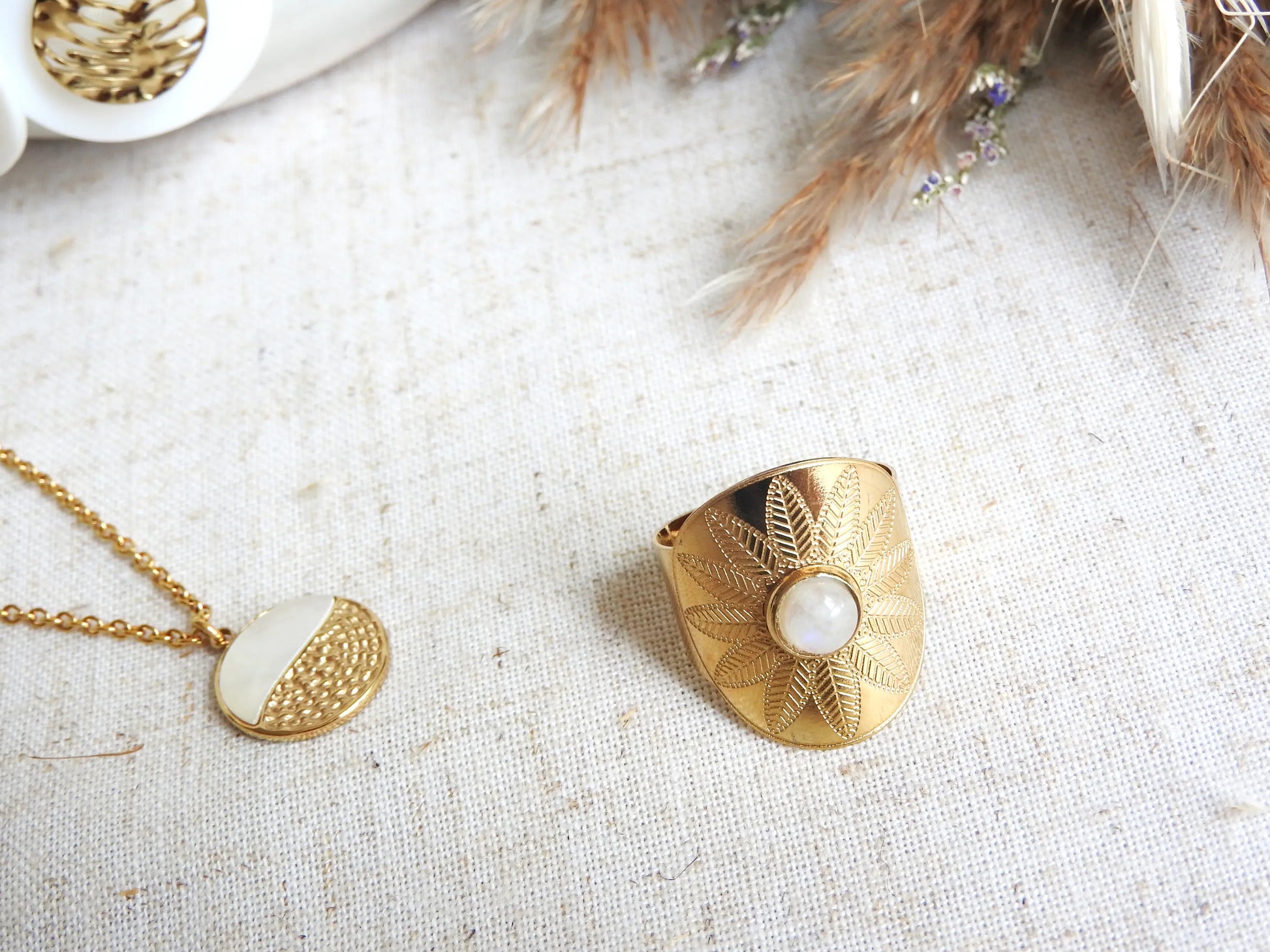 bague style bohème, ethnique réglable en acier inoxydable doré, ornée d'une pierre de lune. Bijou posé sur une nappe beige, à ôté d'un collier à pendentif médaille mi-nacre blanche et mi-texture.