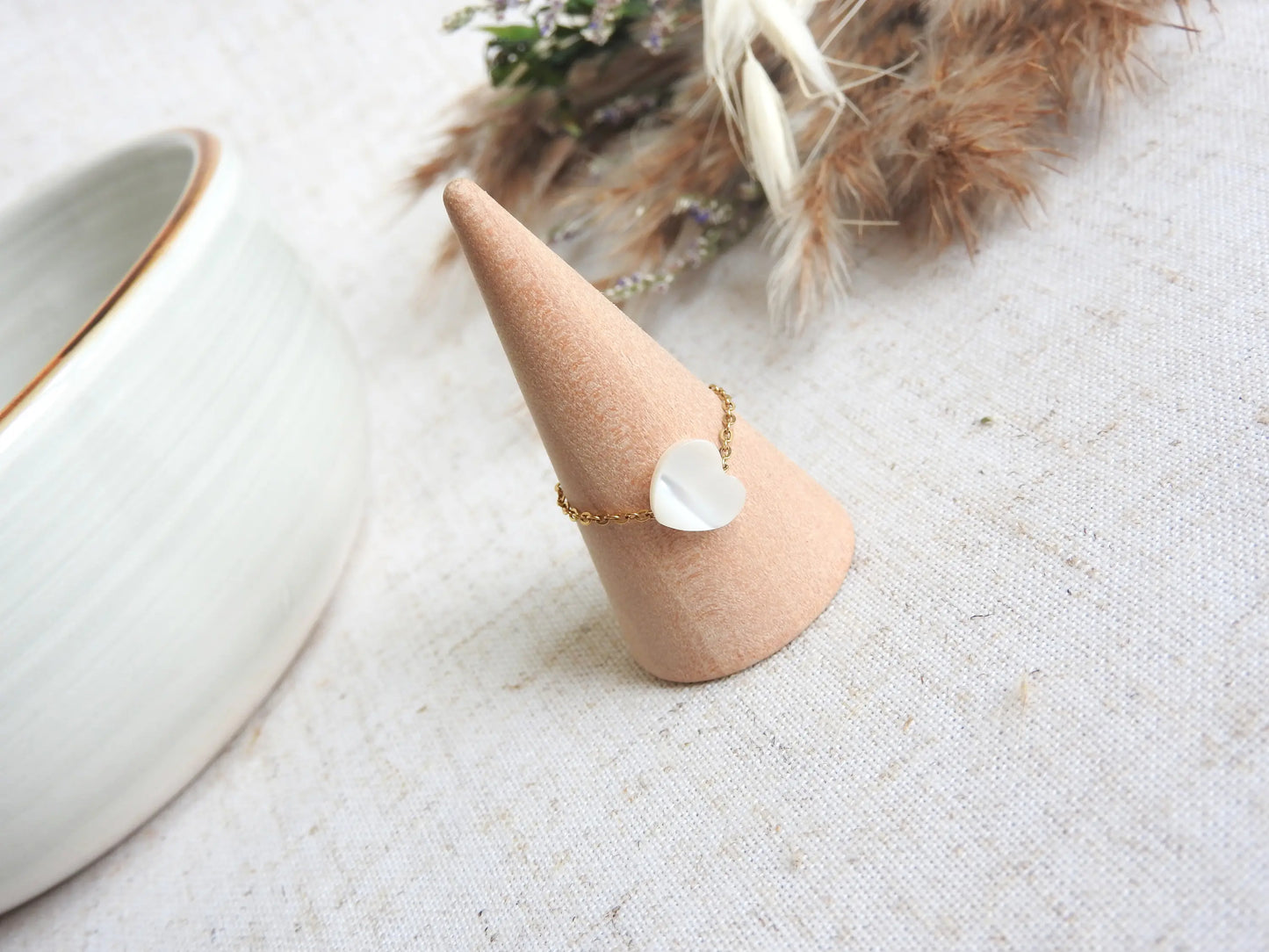 bague chaine en acier inoxydable doré ornée d'une perle coeur en nacre blanche. Bijou posé sur un cône en bambou posé sur une nappe beige décorée d'un mini-bouquet de fleurs.