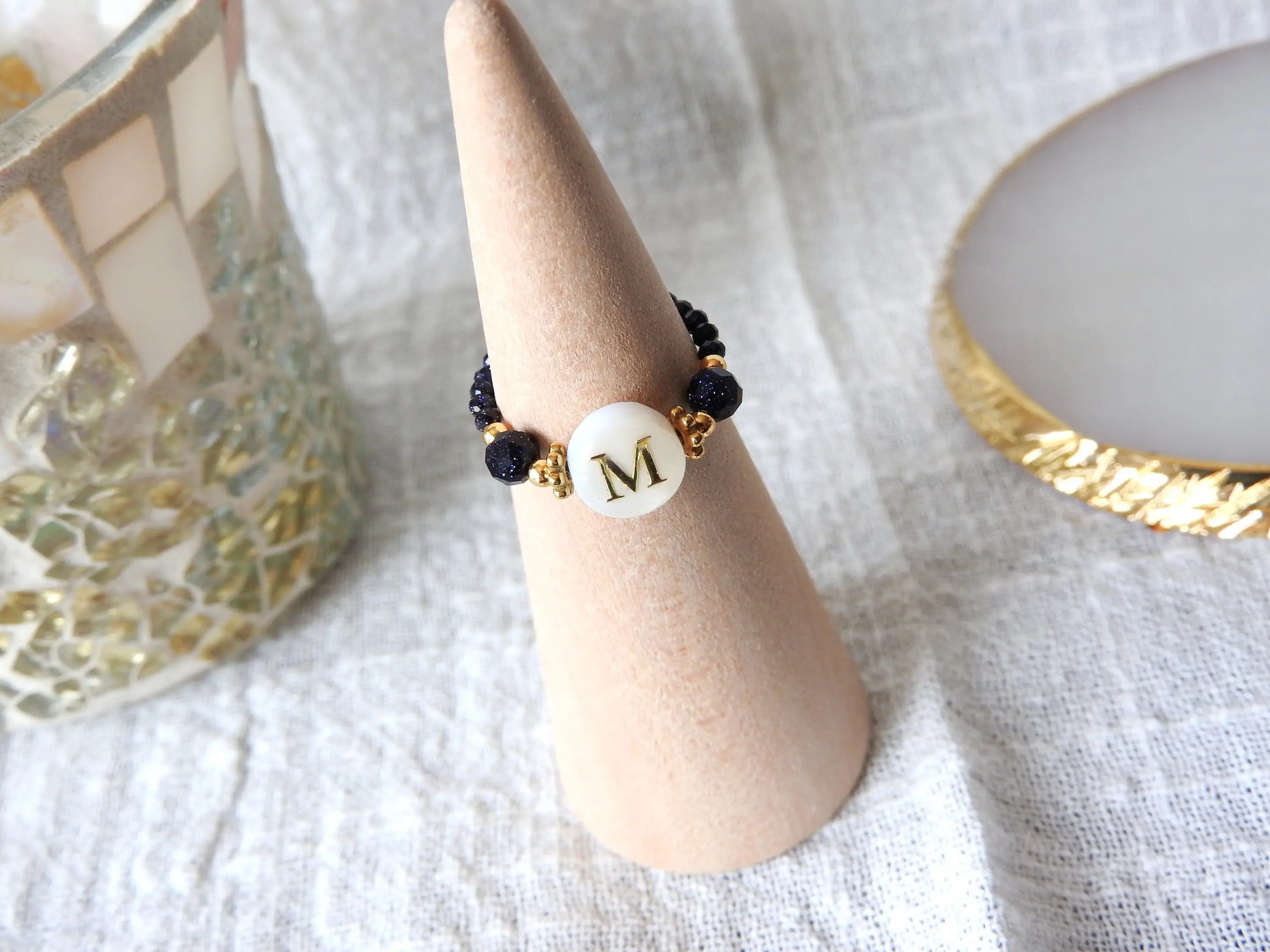 Bague élastique en perles de verre bleu nuit paillette et acier inoxydable doré. Bijou orné d'une perle ronde blanche avec pour motif lettre initiale M doré. Bijou présenté sur un cône bambou.