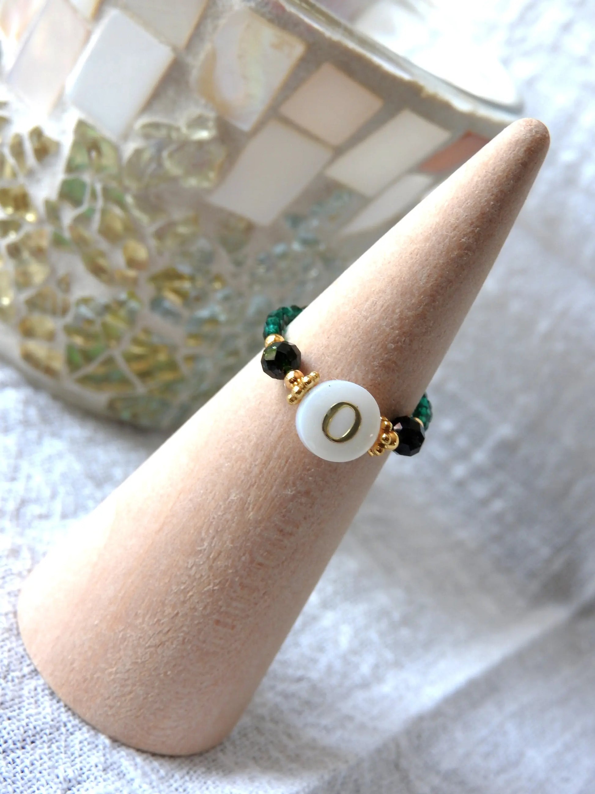 Bague élastique en perles de verre vertes et acier inoxydable doré. Bijou orné d'une perle ronde blanche avec pour motif lettre initiale O doré. Bijou présenté sur un cône bambou.