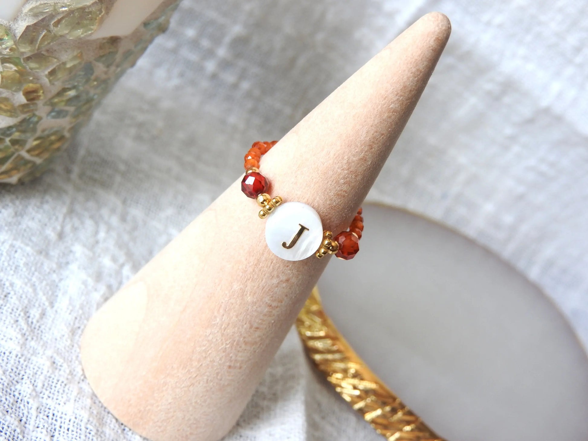 Bague élastique en perles de verre oranges et acier inoxydable doré. Bijou orné d'une perle ronde blanche avec pour motif lettre initiale J doré. Bijou présenté sur un cône bambou.