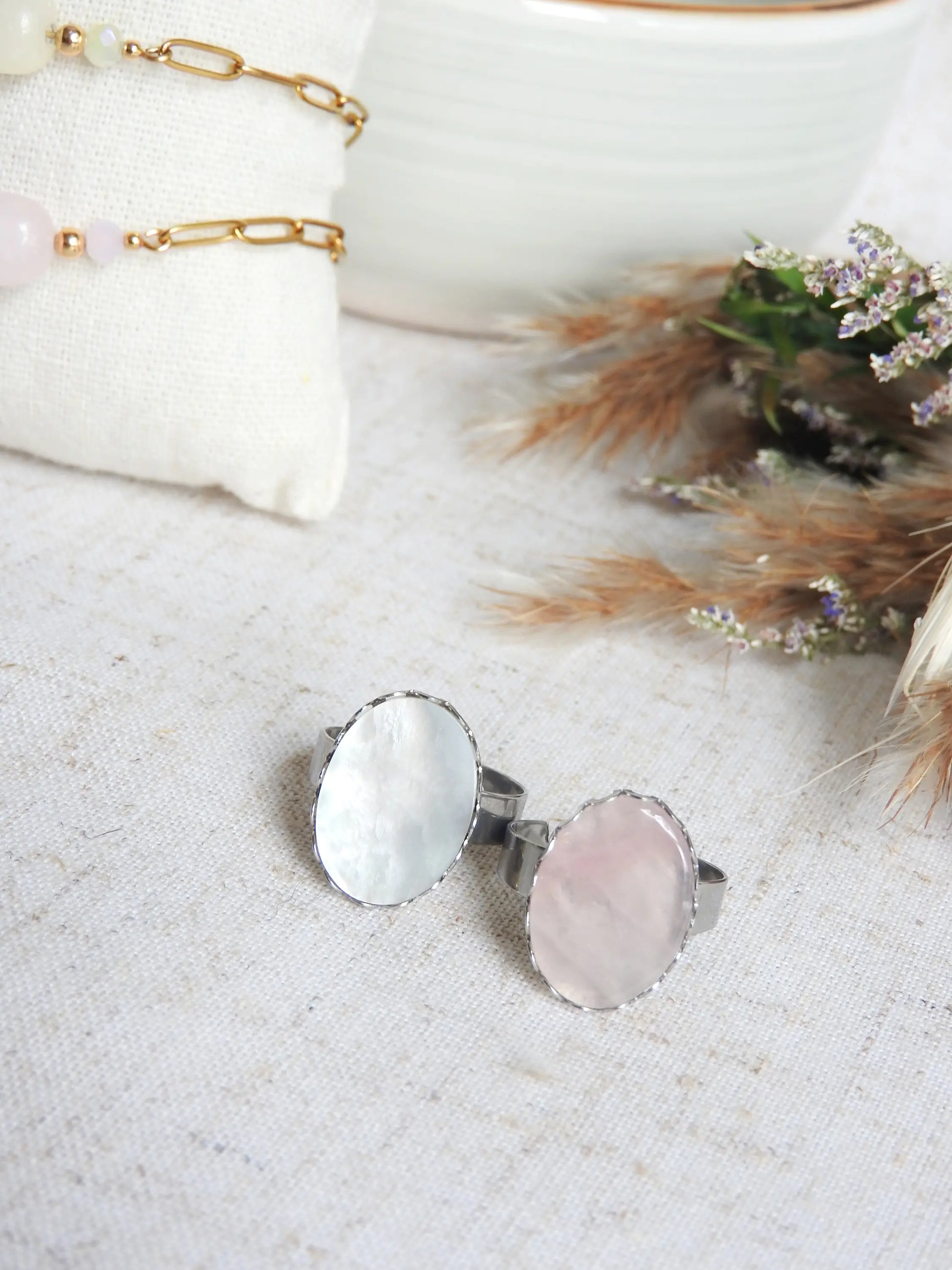 bagues à anneau réglable en acier inoxydable argenté posées à plat sur une nappe beige décorée d'un mini-bouquet de fleurs. Bijoux ornés de piettes naturelles, une pierre plate en nacre blanche et une pierre en quartz rose.