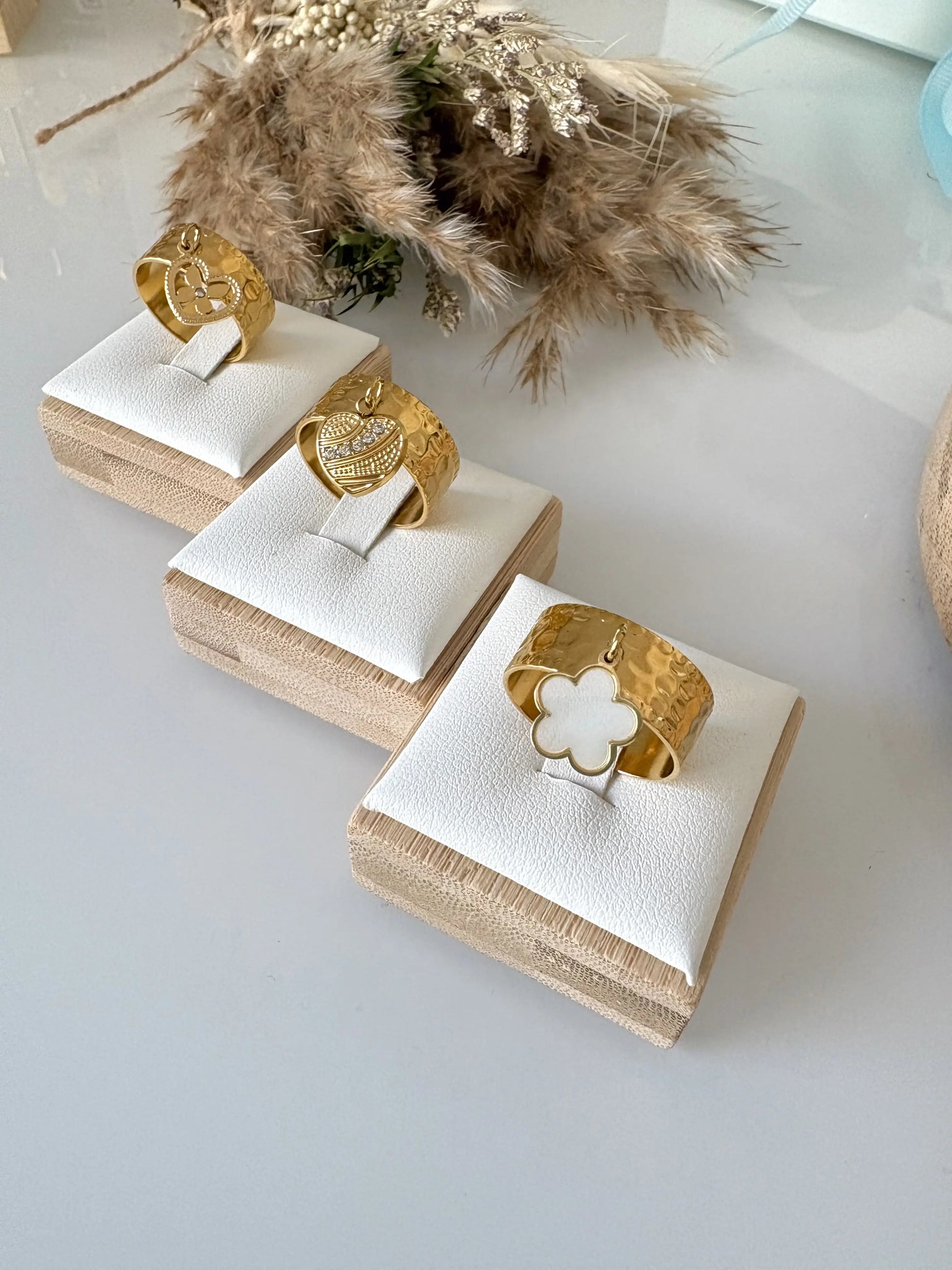 bagues à anneau réglable breloques en acier inoxydable doré, disposées sur des supports individuels en bambou sur une table en verre décorée d'un mini-bouquet de fleurs. Breloque fleur en nacre blanche, breloque coeur ornée de pierre en oxydes dezirconium et breloque coeur papillon strass.