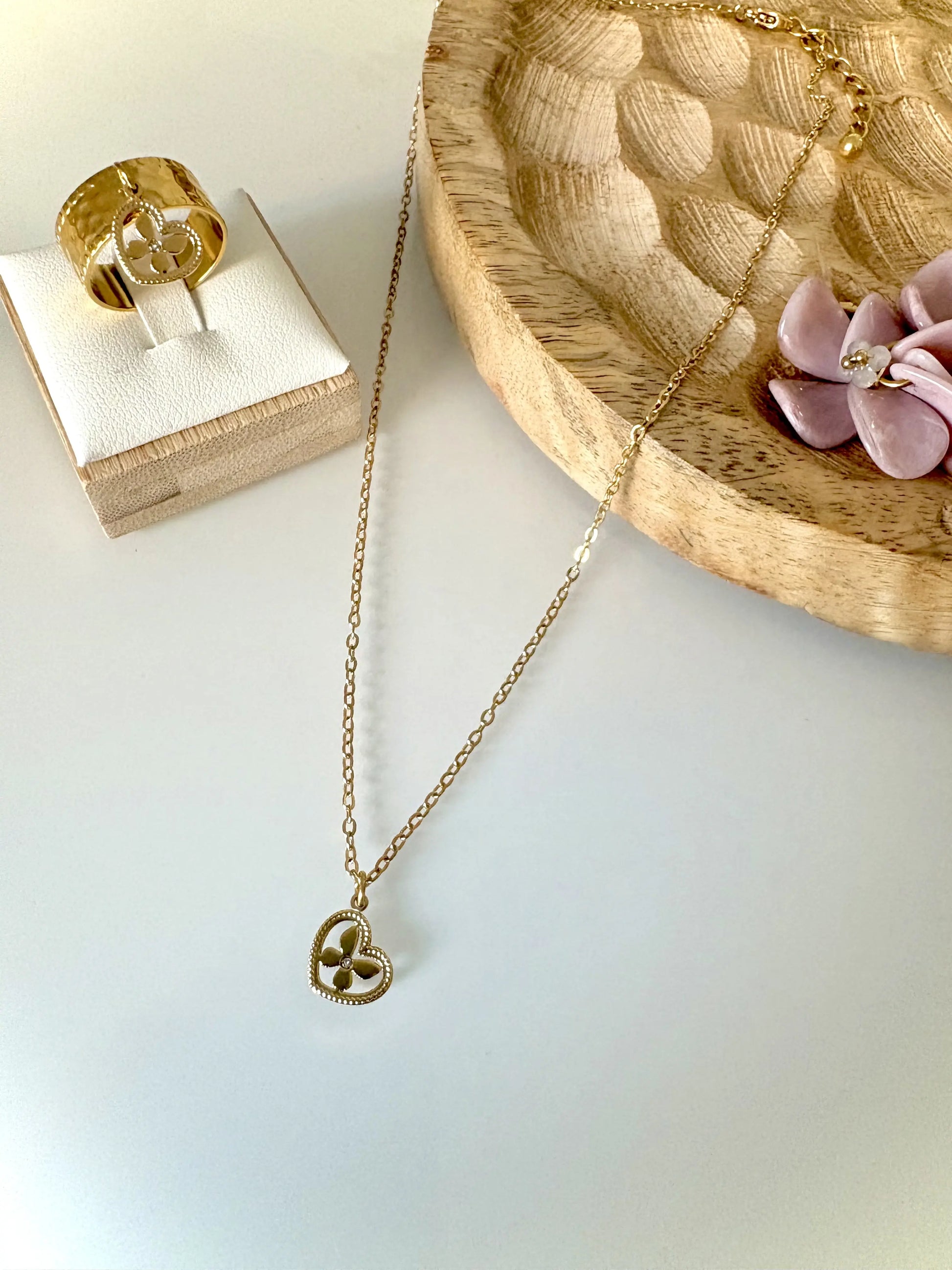 Collier à chaîne fine orné d'un pendentif coeur imbriqué d'un papillon décoré d'un strass en son centre. Bague à anneau martelé à breloque coeur papillon strass en acier inoxydable doré assortie. Bijoux posés sur support bambou.
