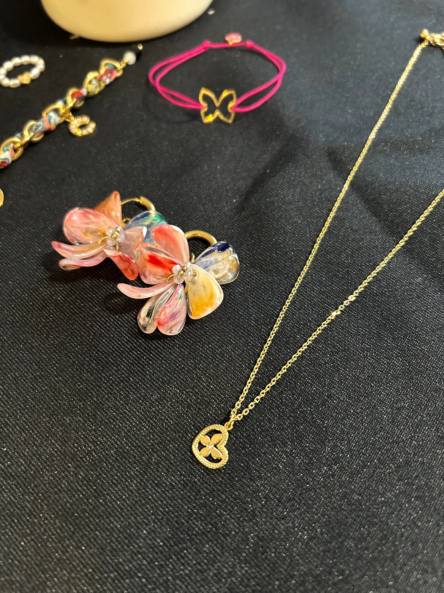 Collier à chaîne fine orné d'un pendentif cœur imbriqué d'un papillon décoré d'un strass en son centre. Bijou en acier inoxydable doré. Boucles d'oreilles mini-créoles à pétales rose fuschia et bracelet cordon fuschia posés à côté du collier. Bijoux posés sur une nappe noire.