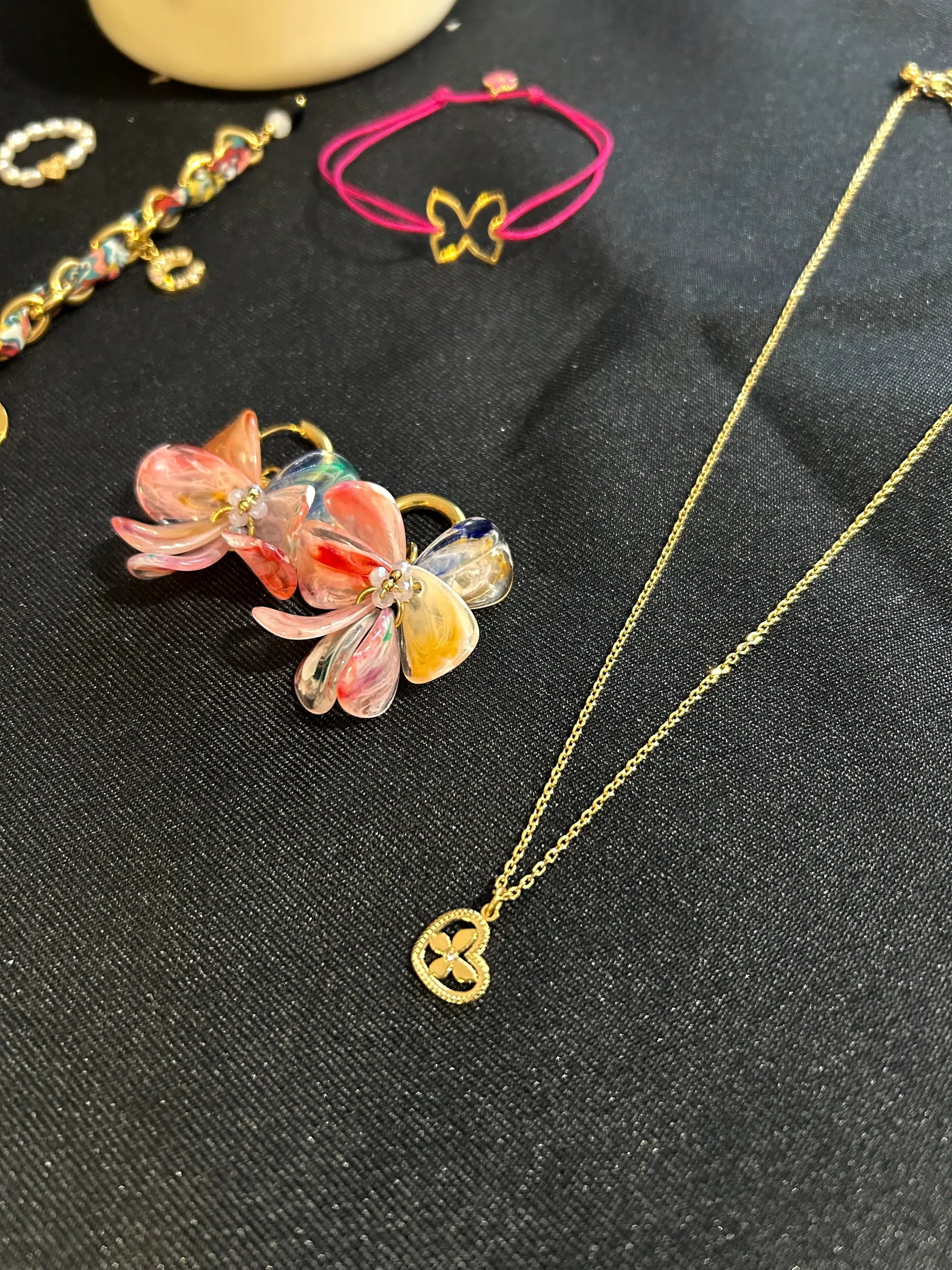 Collier à chaîne fine orné d'un pendentif cœur imbriqué d'un papillon décoré d'un strass en son centre. Bijou en acier inoxydable doré. Boucles d'oreilles mini-créoles à pétales rose fuschia et bracelet cordon fuschia posés à côté du collier. Bijoux posés sur une nappe noire.