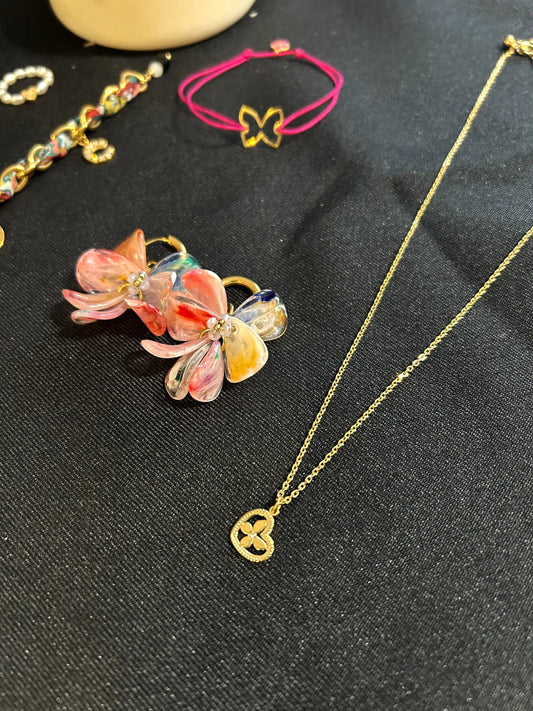 Collier à chaîne fine orné d'un pendentif cœur imbriqué d'un papillon décoré d'un strass en son centre. Bijou en acier inoxydable doré. Boucles d'oreilles mini-créoles à pétales rose fuschia et bracelet cordon fuschia posés à côté du collier. Bijoux posés sur une nappe noire.