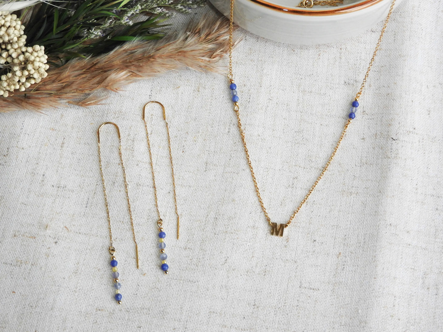 boucles d'oreilles à chaînette traversante en acier inoxydable doré, ornées de perles de verre de tons bleus et jaune. Bijou posé à plat à côté d'un collier assorti à chaîne fine avec une lettre intercalaire en acier inoxydable doré et ornées de perles de verre de tons bleus et jaune.