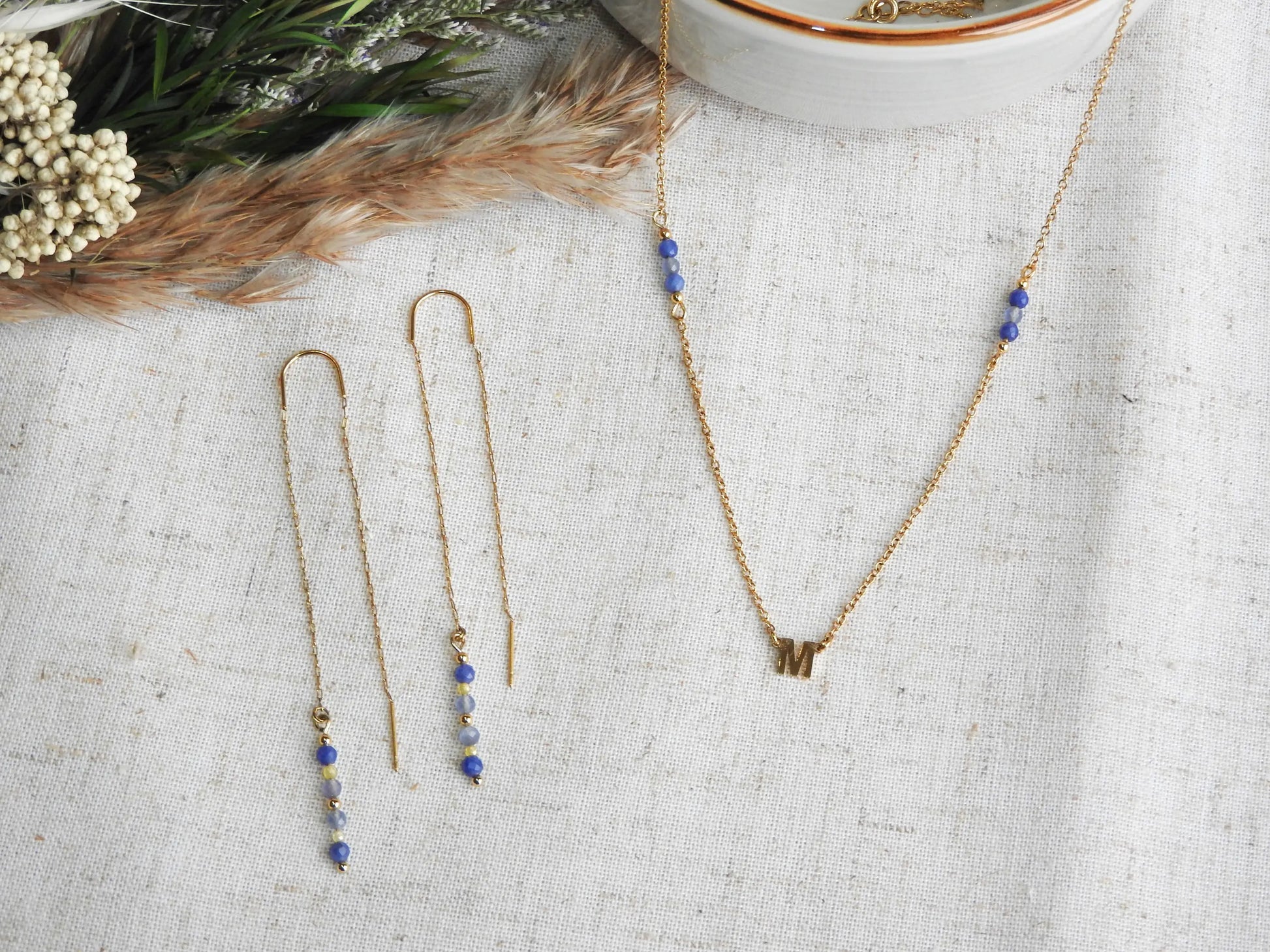 boucles d'oreilles à chaînette traversante en acier inoxydable doré, ornées de perles de verre de tons bleus et jaune. Bijou posé à plat à côté d'un collier assorti à chaîne fine avec une lettre intercalaire en acier inoxydable doré et ornées de perles de verre de tons bleus et jaune.