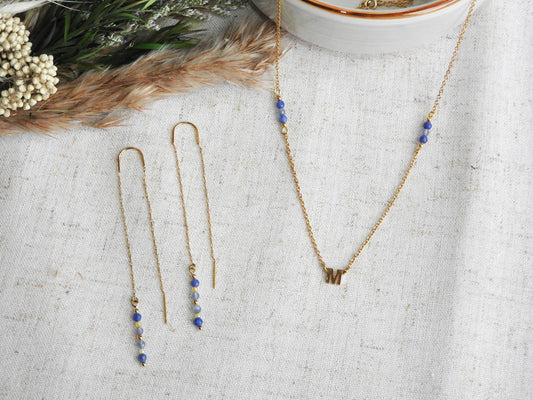 boucles d'oreilles à chaînette traversante en acier inoxydable doré, ornées de perles de verre de tons bleus et jaune. Bijou posé à plat à côté d'un collier assorti à chaîne fine avec une lettre intercalaire en acier inoxydable doré et ornées de perles de verre de tons bleus et jaune.
