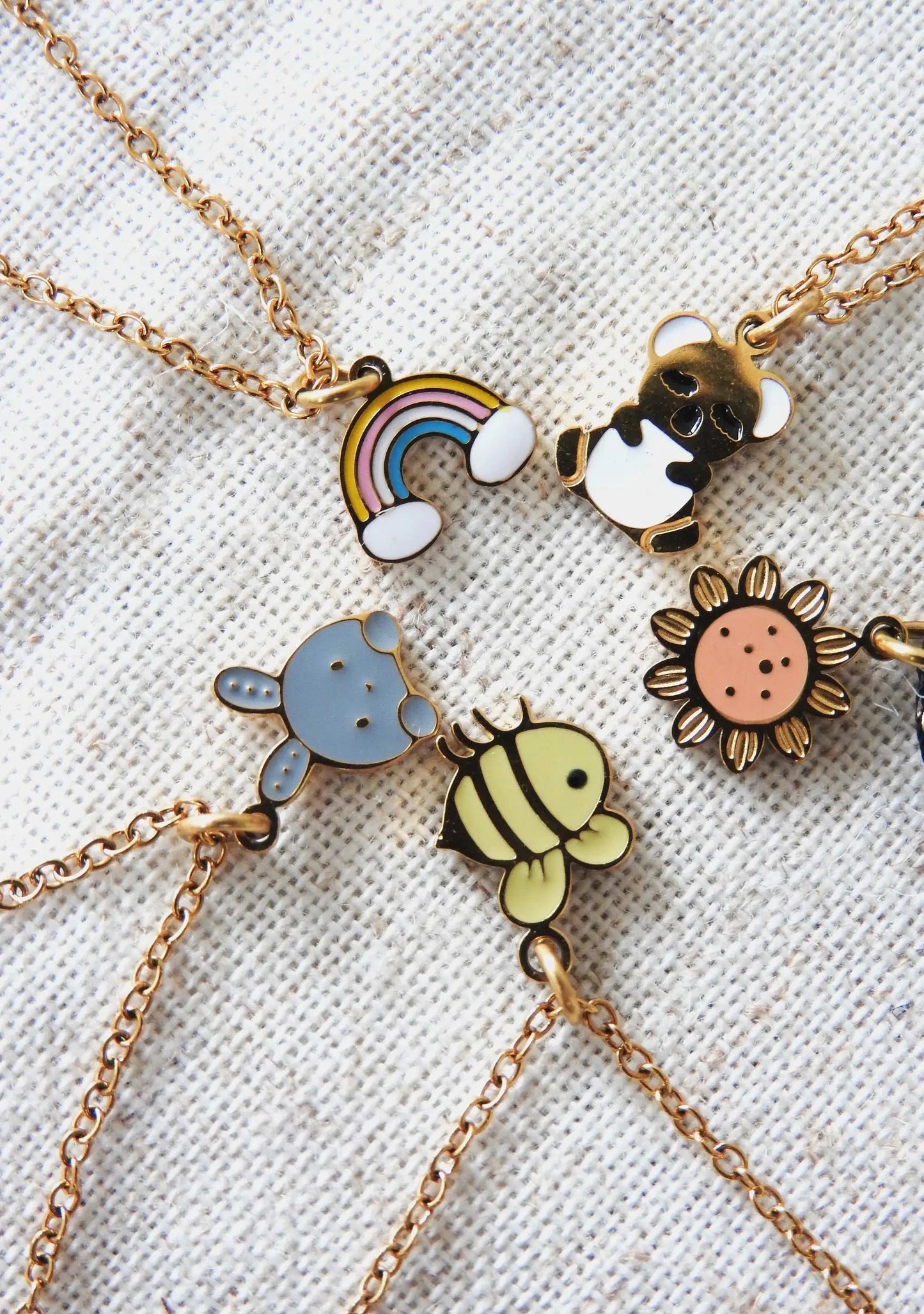 Colliers chaîne pour enfants rangés en cercle en résine époxy colorée et acier inoxydable doré. Pendentifs koala, arc-en-ciel, tournesol, lapin et abeille. Bijoux posés à plat sur une nappe beige.