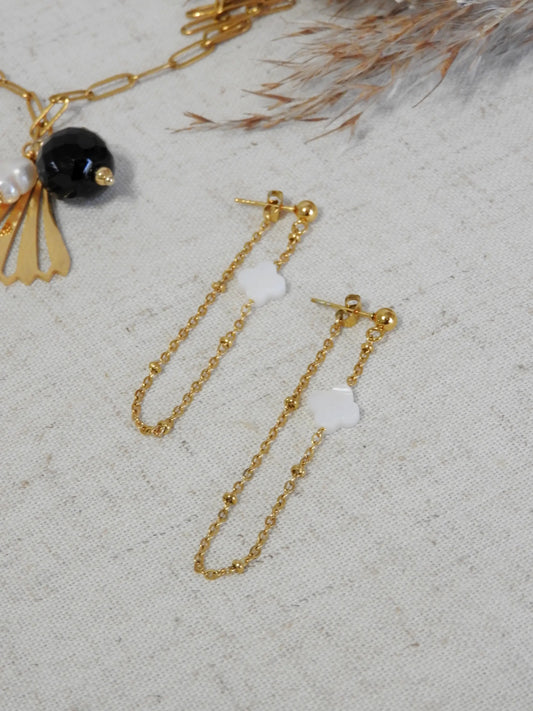 boucles d'oreilles chaînettes en acier inoxydable doré ornées d'une perle trèfle en nacre blanche. Bijou posé à plat sur une nappe beige décorée d'une brindille et à un collier avec une perle d'eau douce et onyx noir.