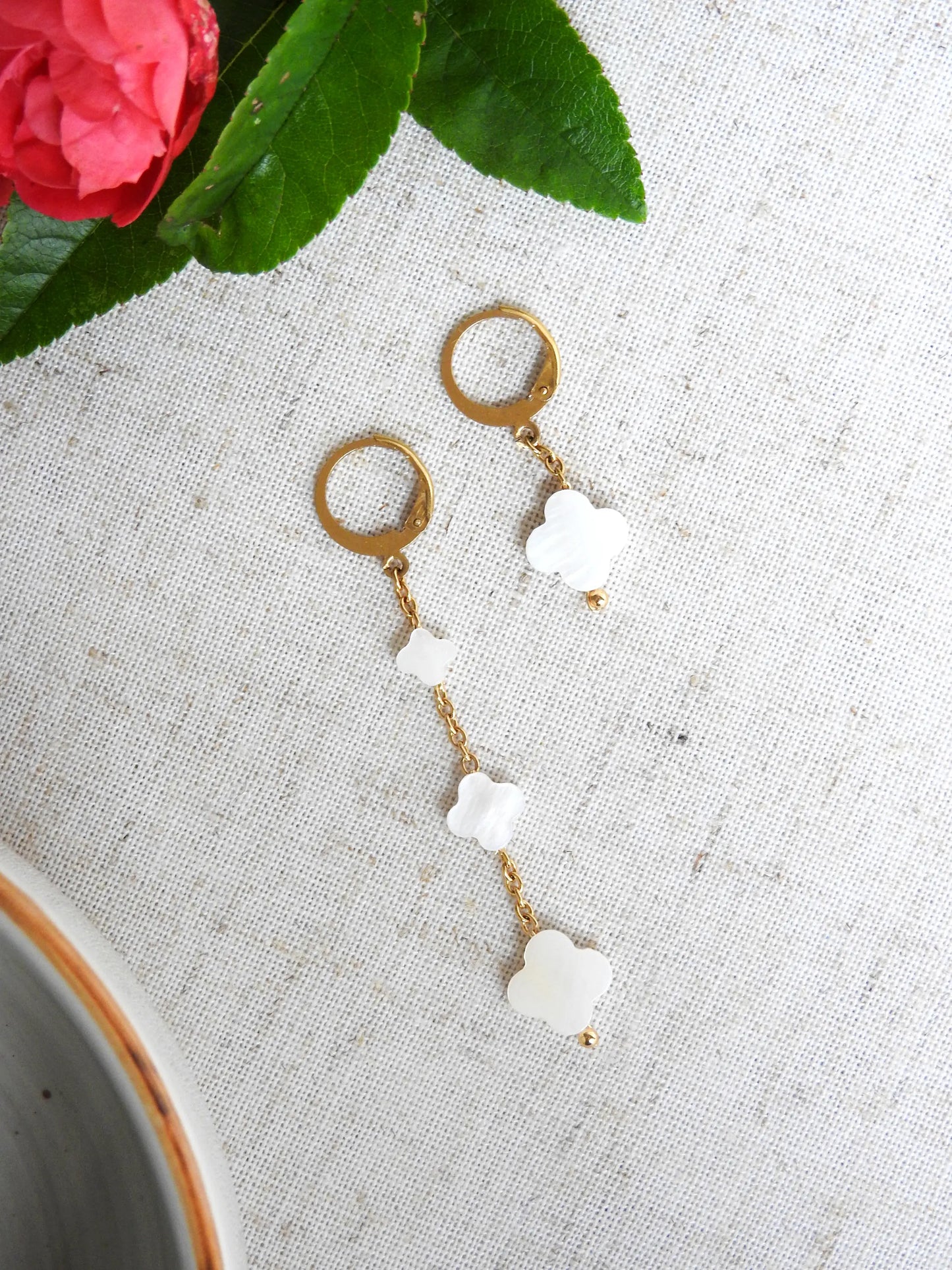 boucles d'oreilles à chaînes pendantes asymétriques ornées de perles trèfle en nacre blanche. Bijou posé à plat sur une nappe blanche décorée de roses.