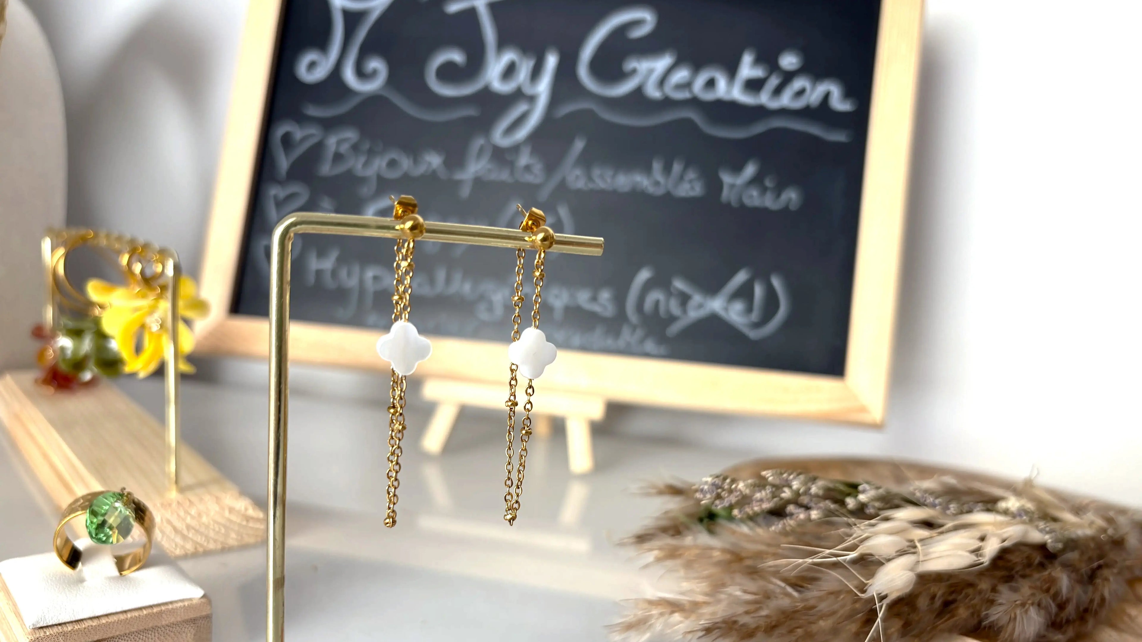 Boucles d'oreilles chaînette perlée en acier inoxydable doré, ornées d'une perle trèfle en nacre blanche. Bijou suspendu à un support métal doré et bambou. Arrière plan des boucles d'oreilles en pétales en acrylique coloré jaune, vert olive et ambre. Bague anneau avec breloque goutte verte en oxyde de zirconium. Bijoux posés sur table en verre décoré d'un plateau bambou décoré de brindilles.