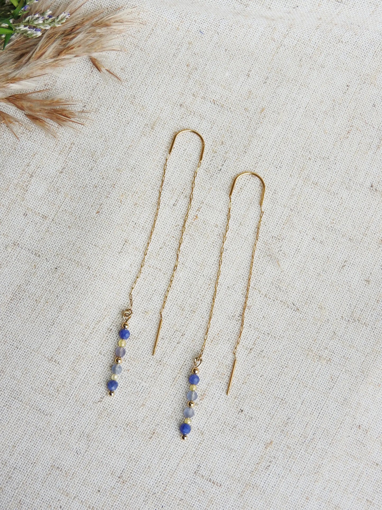 boucles d'oreilles à chaînette traversante en acier inoxydable doré, posé à plat sur une nappe beige en coton. Bijou orné de perles de verre de tons bleus et jaune.