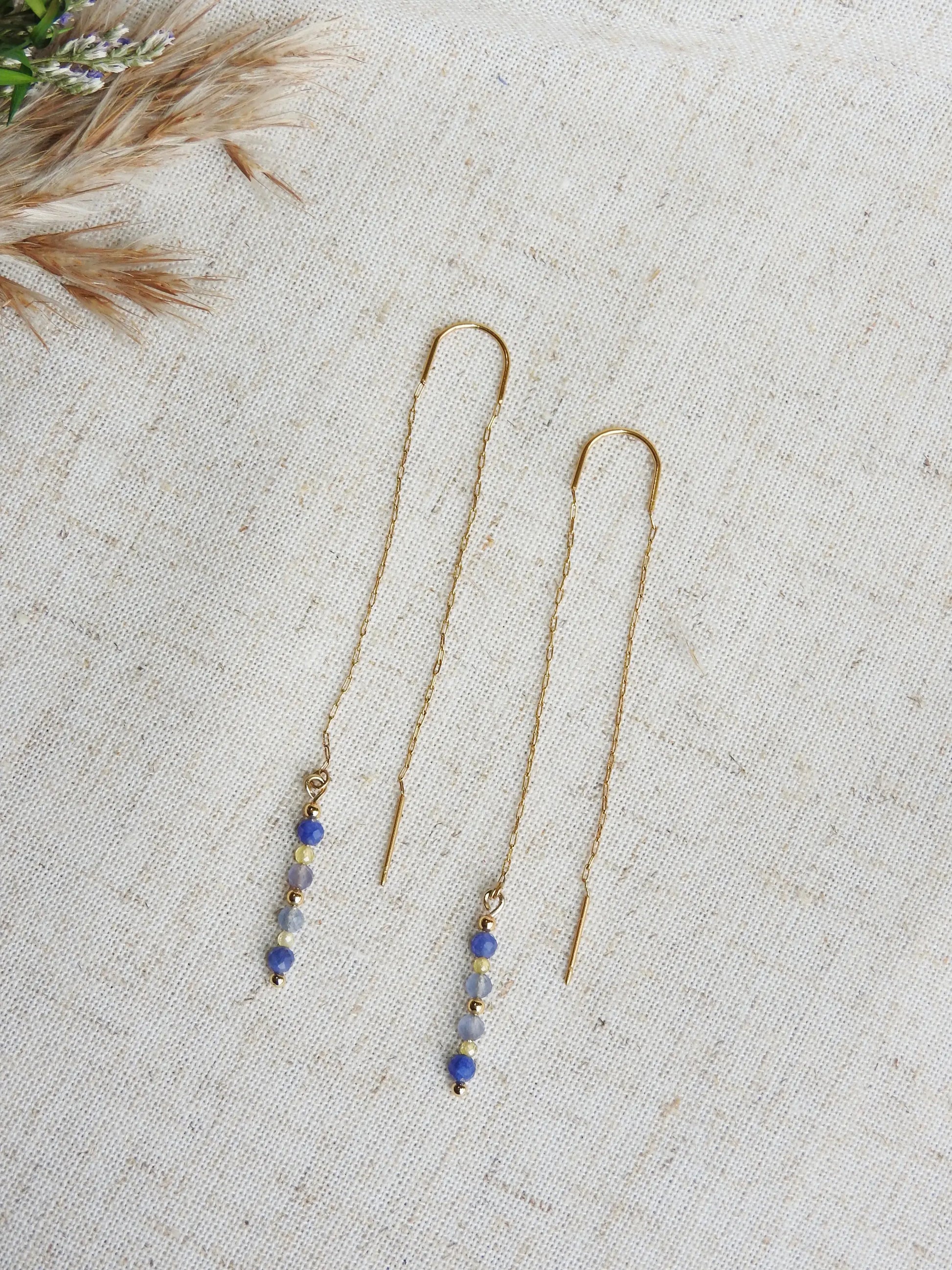 boucles d'oreilles à chaînette traversante en acier inoxydable doré, posé à plat sur une nappe beige en coton. Bijou orné de perles de verre de tons bleus et jaune.