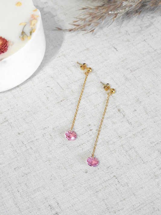 Boucles d'oreilles chaînettes pendantes en acier inoxydable doré, orné d'une pierre cristal rose suspendue. Bijou posé à plat sur une nappe beige décorée d'une bougie fleurie.