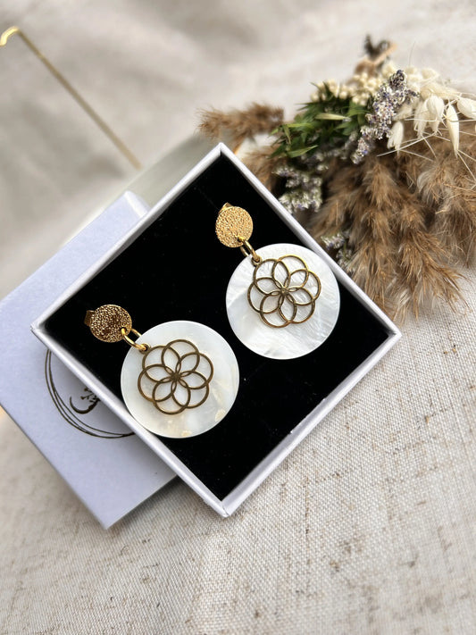 Boucles d'oreilles clous à disque martelé rangées dans un écrin blanc avec une mousse noire. Bijou orné d'un sequin en nacre blanche superposée d'une fleur évidée en acier inoxydable doré.