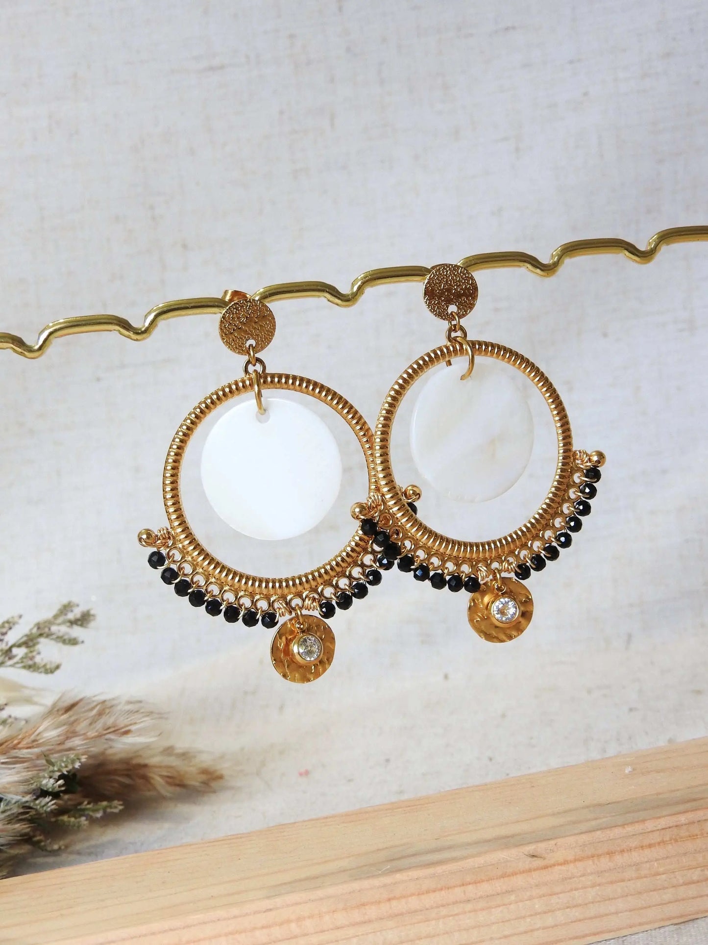 boucles d'oreilles clous à disque martelé ornées d'un chandelier décoré d'un double sequin en nacre blanche et acier inoxydable doré et des perles de verre noires en décoration. Finition de la boucle avec une pierre de naissance cristal pour le mois d'avril. Bijoux suspendus à une barre métallique doré.