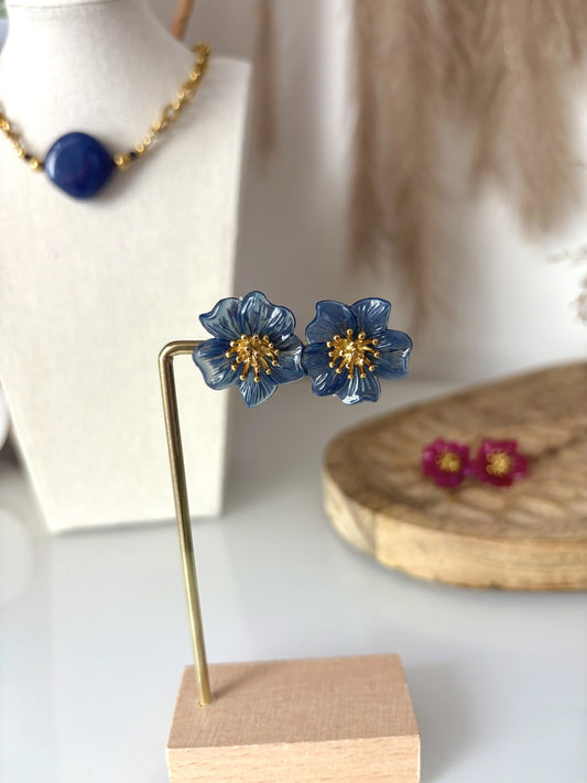 boucles d'oreilles clous à pistil en acier inoxydable doré et orné d'une fleur hibiscus bleu foncé. Bijoux présentés sur un présentoir métallique doré et bambou avec en fond un collier à grosse perle losange bleu foncé, posé sur un buste présentoir.
