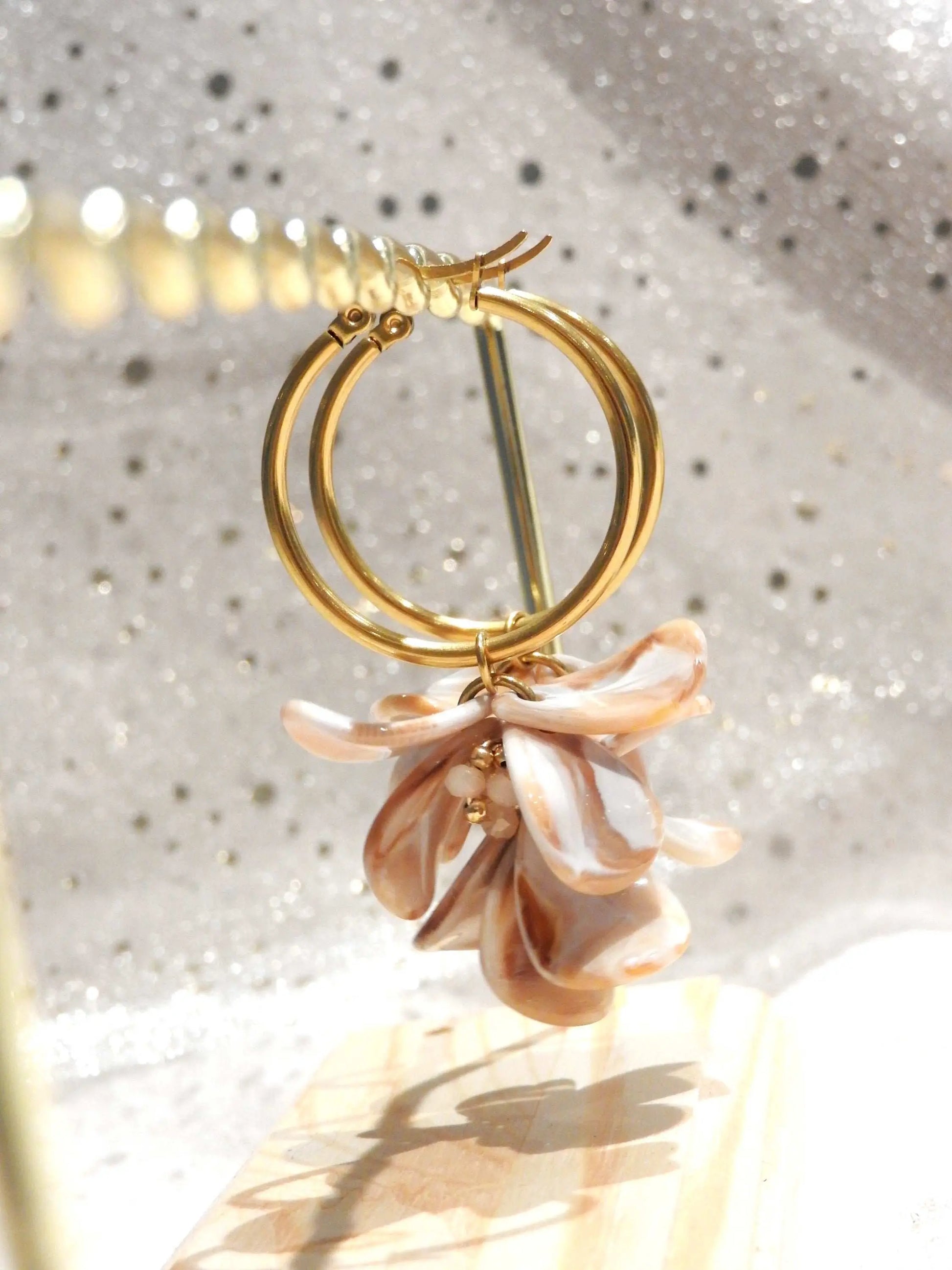 Boucles d'oreilles créoles en acier inoxydable doré suspendues à un support métallique doré et en bambou posé sur une nappe beige. Boucles décorées de pétales colorées beige à effet marbré, formant un bouquet floral suspendu à la créole.