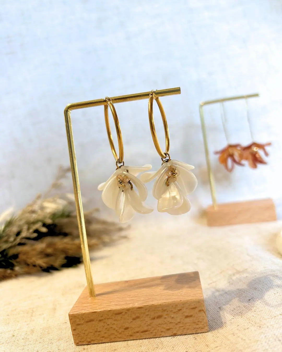 Boucles d'oreilles créoles en acier inoxydable doré suspendues à un support métallique doré et en bambou posé sur une nappe beige. Boucles décorées de pétales colorées blanches à effet marbré, formant un bouquet floral suspendu à la créole.