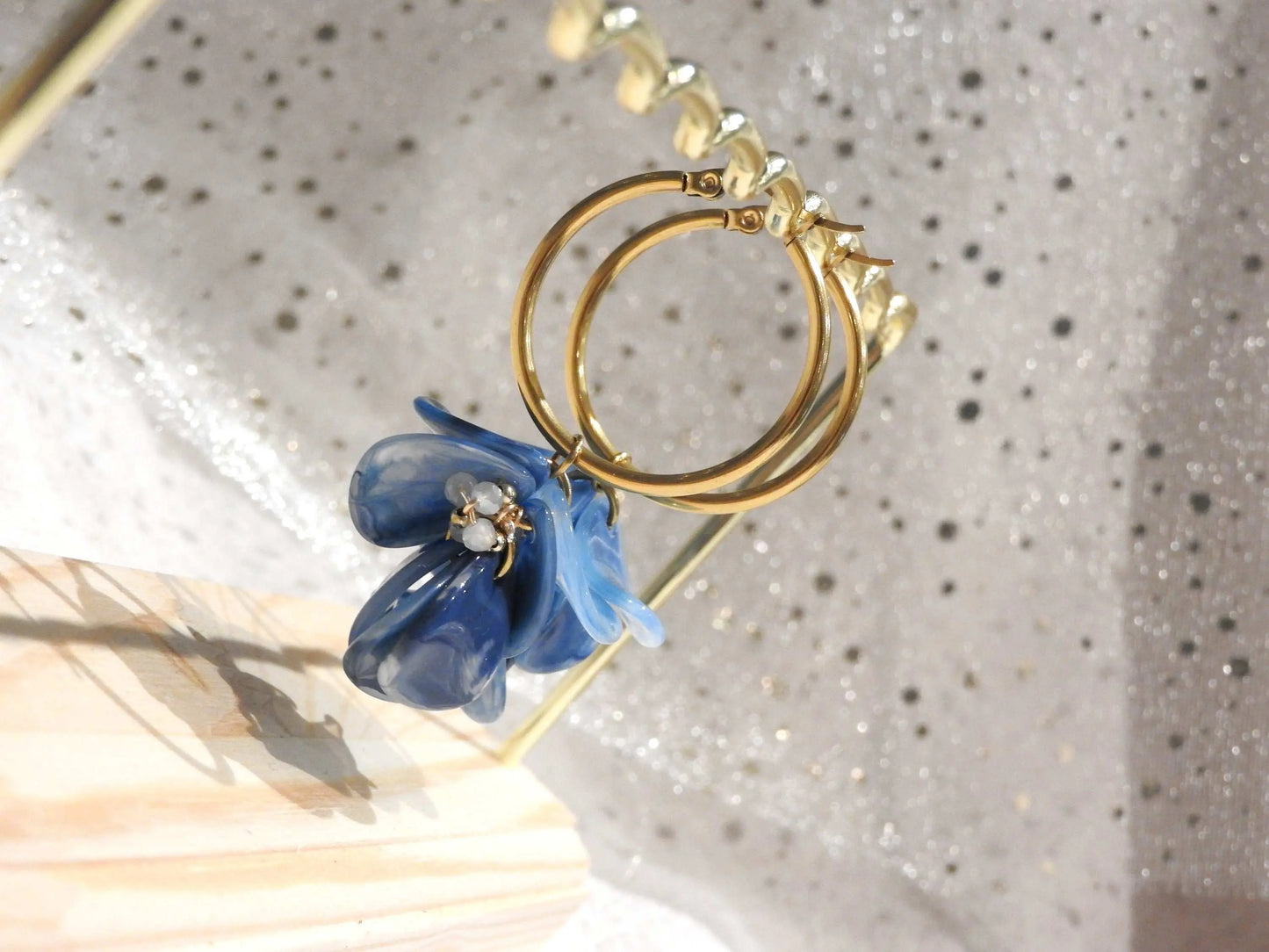 Boucles d'oreilles créoles en acier inoxydable doré suspendues à un support métallique doré et en bambou posé sur une nappe beige. Boucles décorées de pétales colorées bleu à effet marbré, formant un bouquet floral suspendu à la créole.