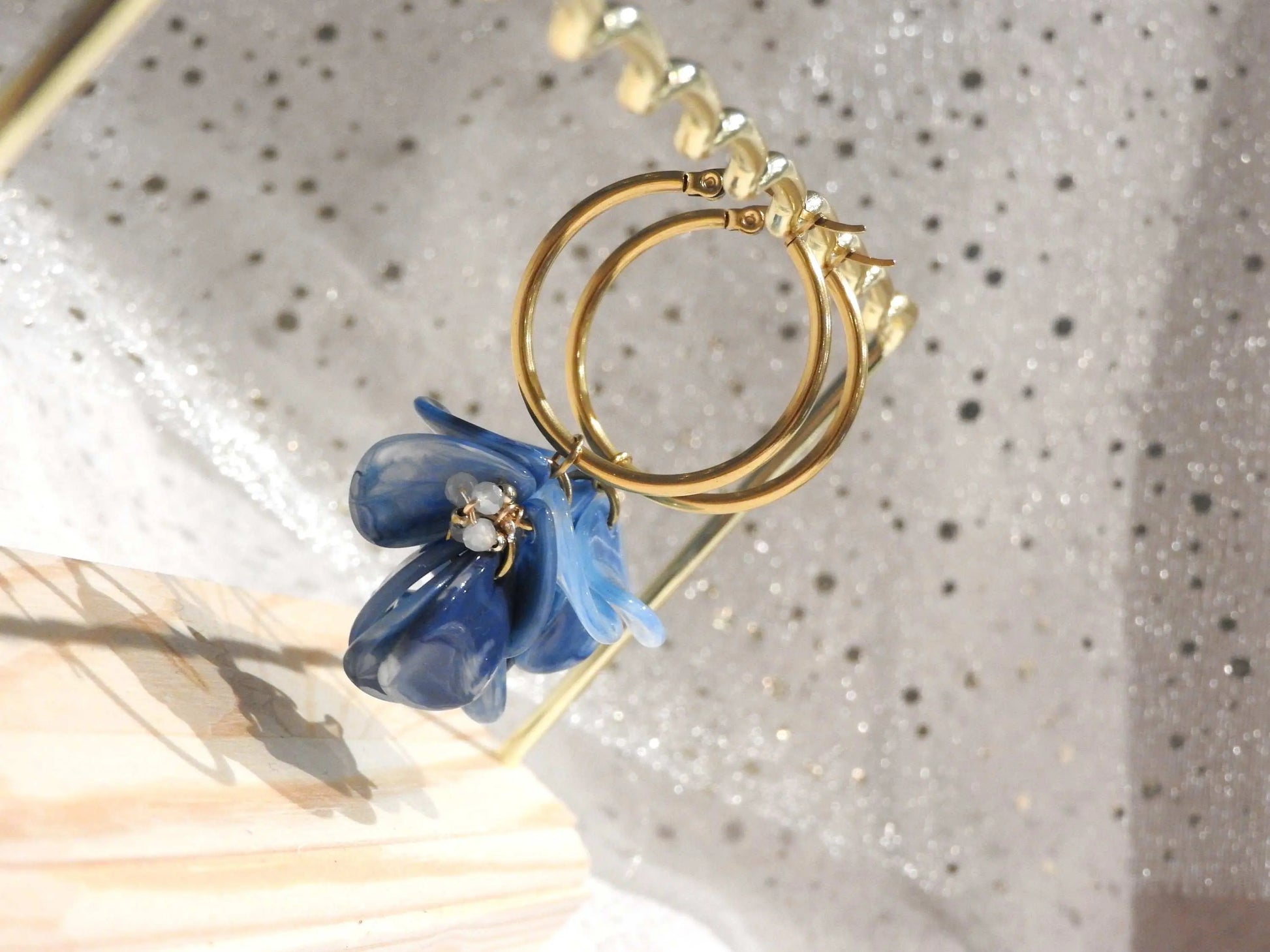 Boucles d'oreilles créoles en acier inoxydable doré suspendues à un support métallique doré et en bambou posé sur une nappe beige. Boucles décorées de pétales colorées bleu à effet marbré, formant un bouquet floral suspendu à la créole.