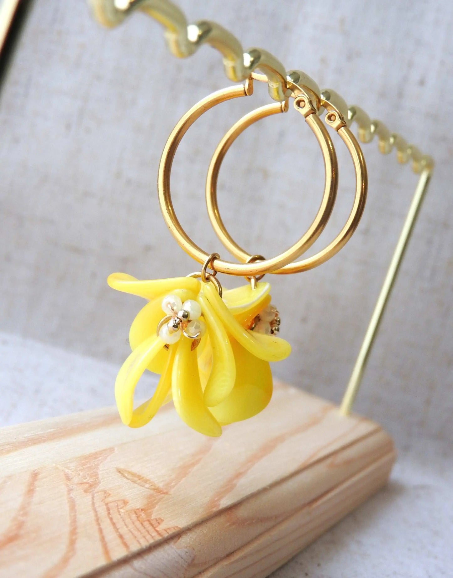 Boucles d'oreilles créoles en acier inoxydable doré suspendues à un support métallique doré et en bambou posé sur une nappe beige. Boucles décorées de pétales colorées jaune à effet marbré, formant un bouquet floral suspendu à la créole.