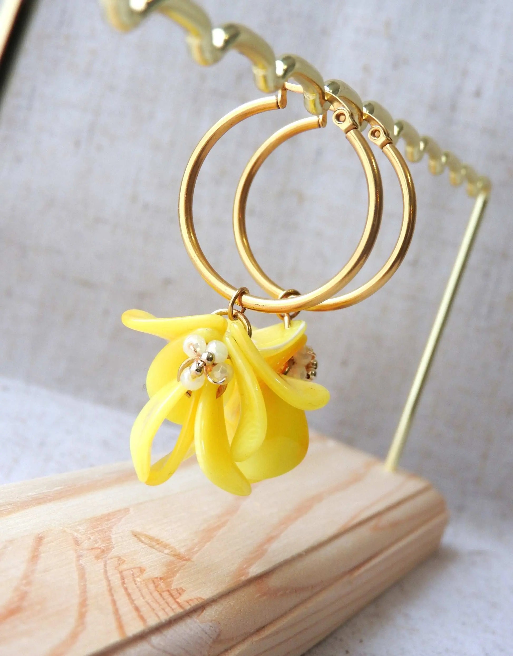 Boucles d'oreilles créoles en acier inoxydable doré suspendues à un support métallique doré et en bambou posé sur une nappe beige. Boucles décorées de pétales colorées jaune à effet marbré, formant un bouquet floral suspendu à la créole.