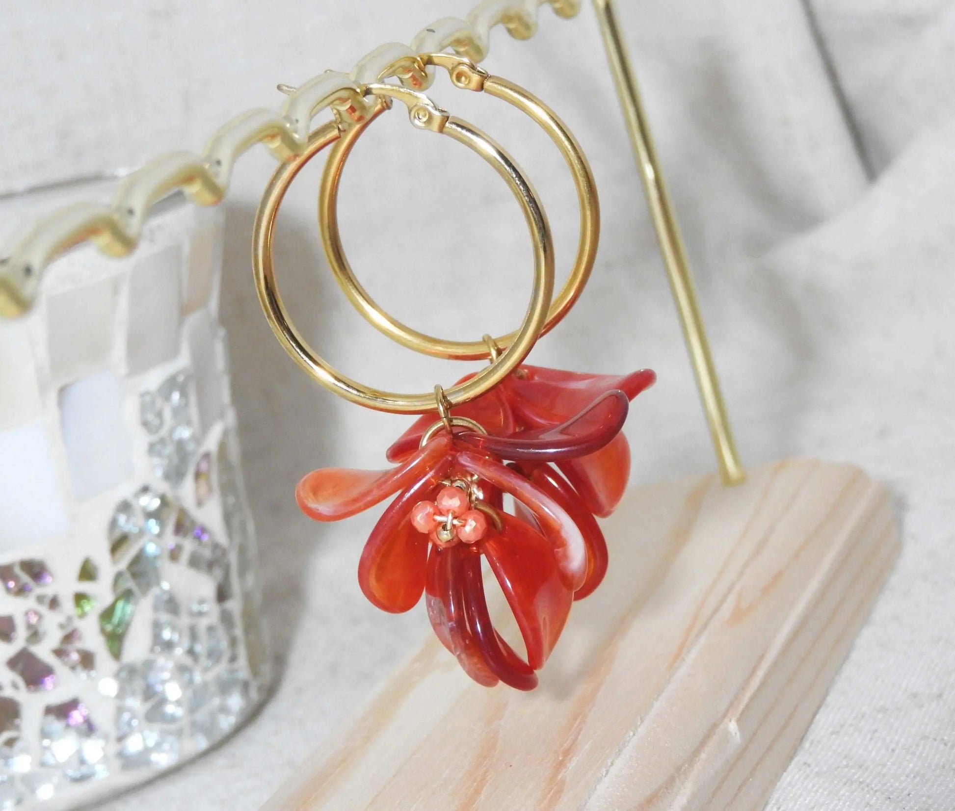 Boucles d'oreilles créoles en acier inoxydable doré suspendues à un support métallique doré et en bambou posé sur une nappe beige. Boucles décorées de pétales colorées rouge à effet marbré, formant un bouquet floral suspendu à la créole.