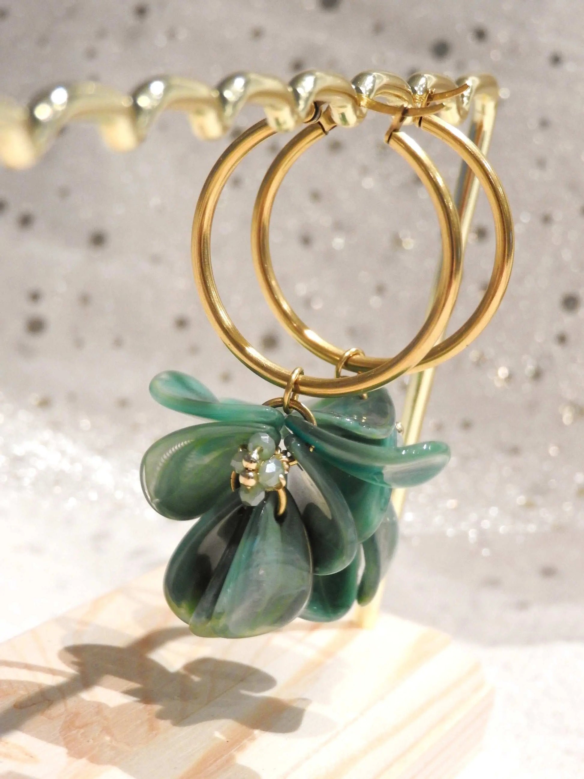 Boucles d'oreilles créoles en acier inoxydable doré suspendues à un support métallique doré et en bambou posé sur une nappe beige. Boucles décorées de pétales colorées vert à effet marbré, formant un bouquet floral suspendu à la créole.