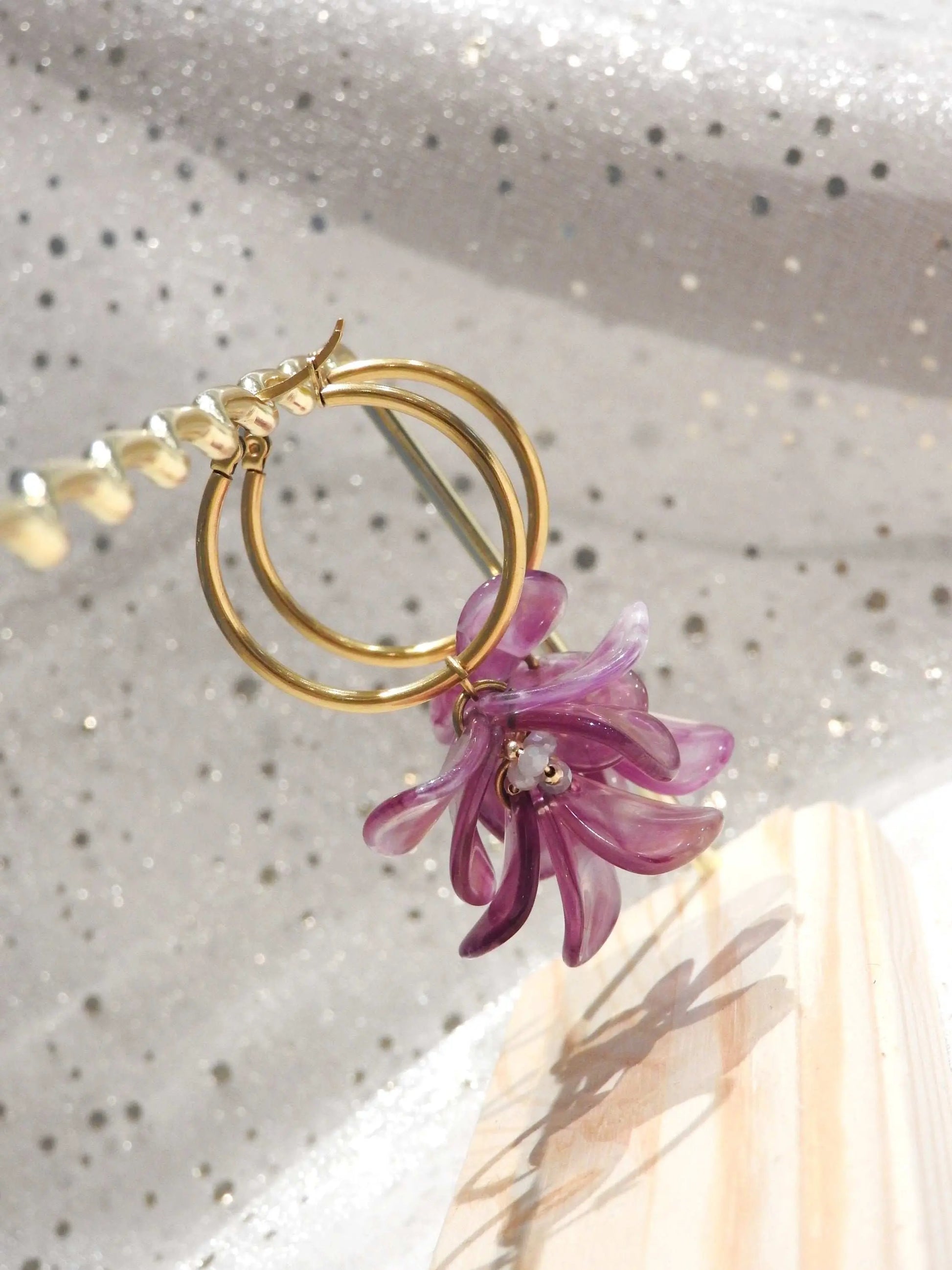 Boucles d'oreilles créoles en acier inoxydable doré suspendues à un support métallique doré et en bambou posé sur une nappe beige. Boucles décorées de pétales colorées violet à effet marbré, formant un bouquet floral suspendu à la créole.