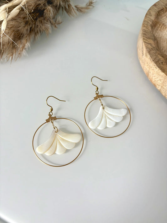 Boucles d'oreilles crochets avec fleur en nacre blanche suspendue et encerclée par une créole en acier inoxydable doré. Bijou posé à plat sur une table en verre décorée d'un plateau en bambou et brindille
