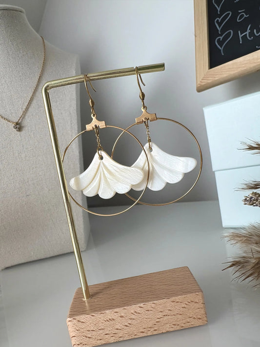 Boucles d'oreilles crochets avec fleur en nacre blanche suspendue et encerclée par une créole en acier inoxydable doré. Bijou présenté sur un support en métal doré et bambou.