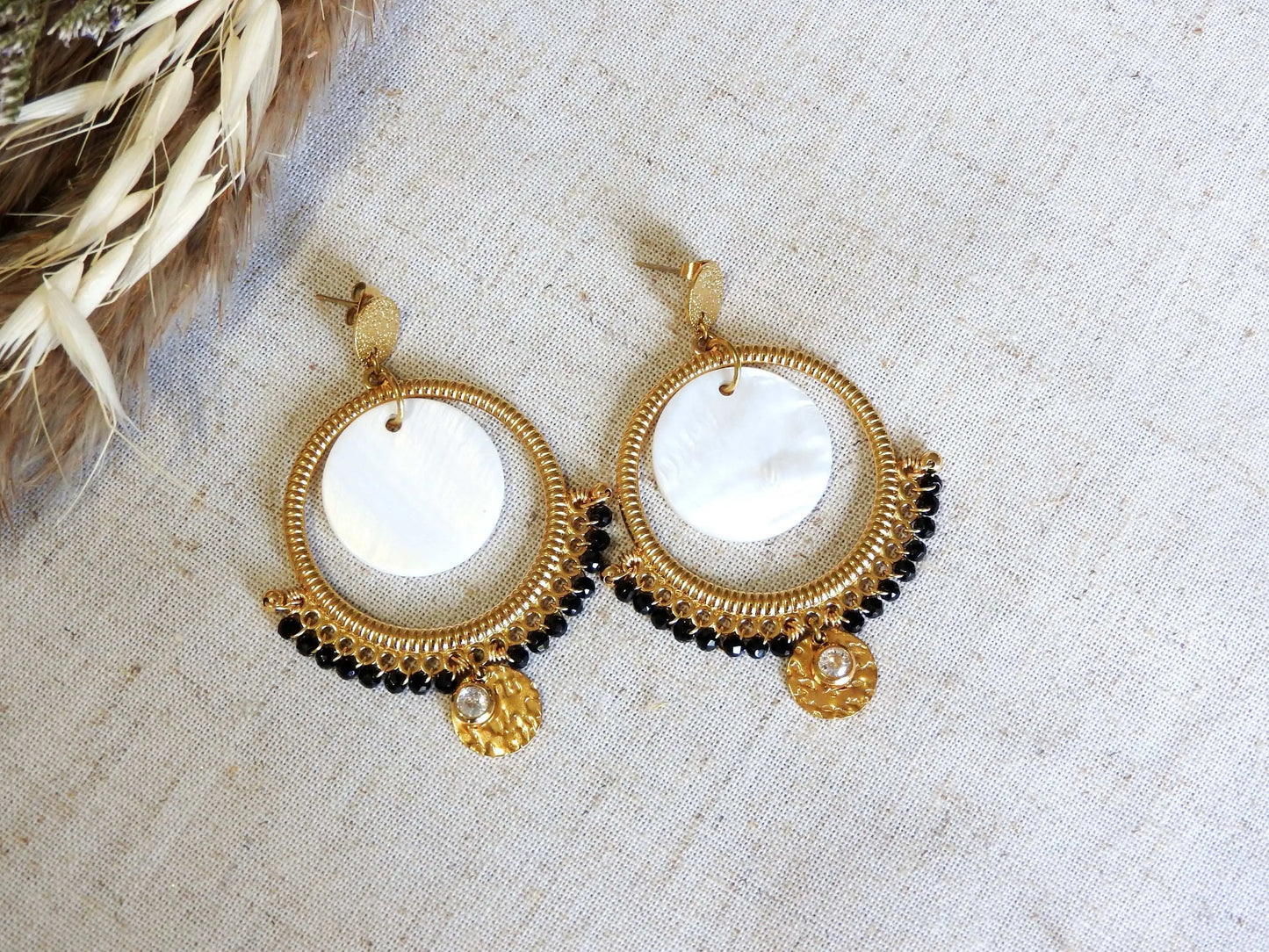 boucles d'oreilles clous à disque martelé ornées d'un chandelier décoré d'un double sequin en nacre blanche et acier inoxydable doré et des perles de verre noires en décoration. Finition de la boucle avec une pierre de naissance cristal pour le mois d'avril. Bijoux suspendus à une barre métallique doré.