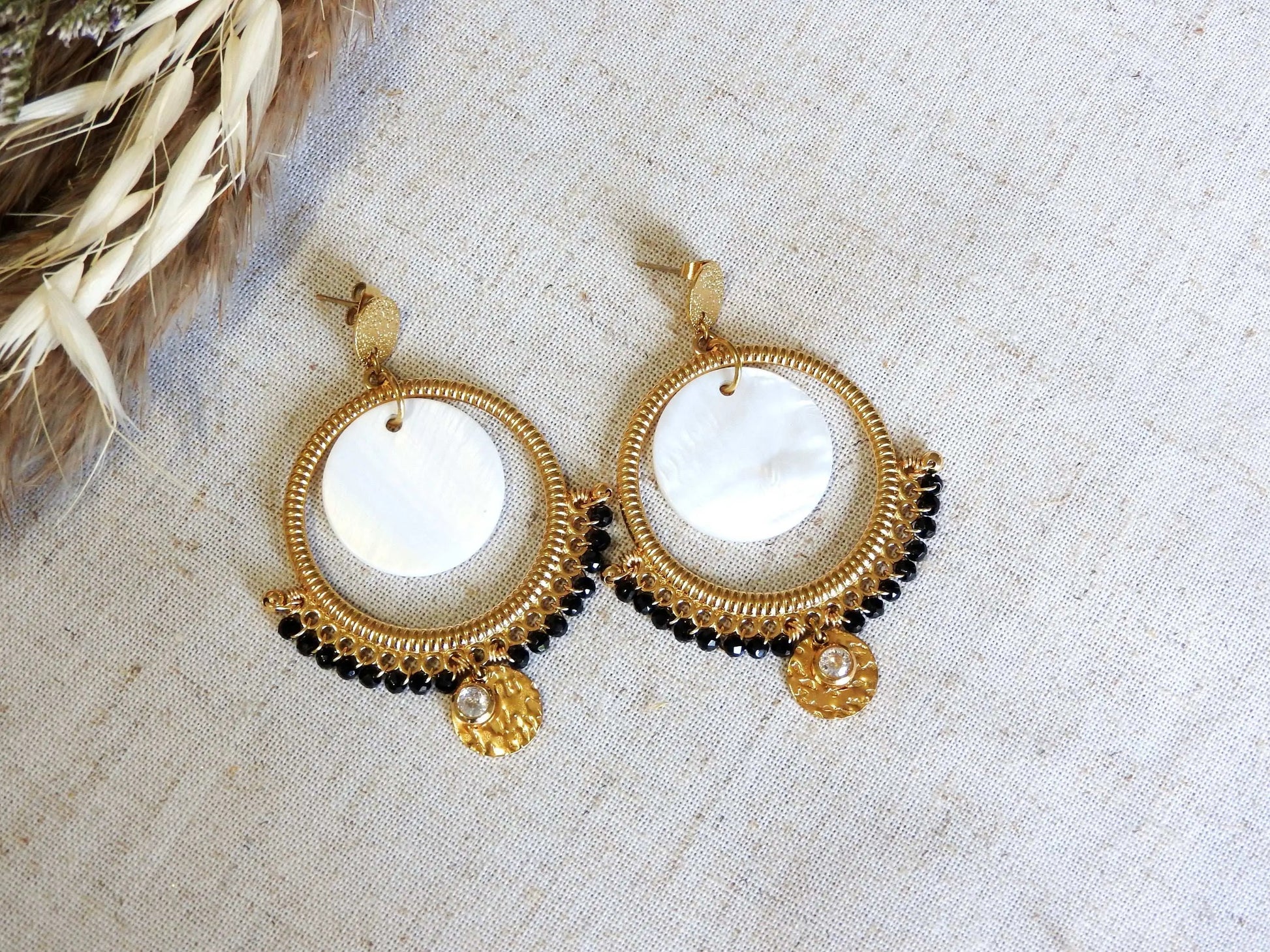 boucles d'oreilles clous à disque martelé ornées d'un chandelier décoré d'un double sequin en nacre blanche et acier inoxydable doré et des perles de verre noires en décoration. Finition de la boucle avec une pierre de naissance cristal pour le mois d'avril. Bijoux suspendus à une barre métallique doré.