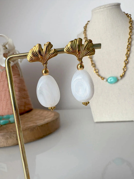 Boucles d'oreilles ornées d'une feuille de ginkgo en acier inoxydable doré avec une grande perle irrégulière blanche suspendue. Bijou présenté sur un support métallique doré posé sur une table avec en fond un buste présentoir présentant un collier.
