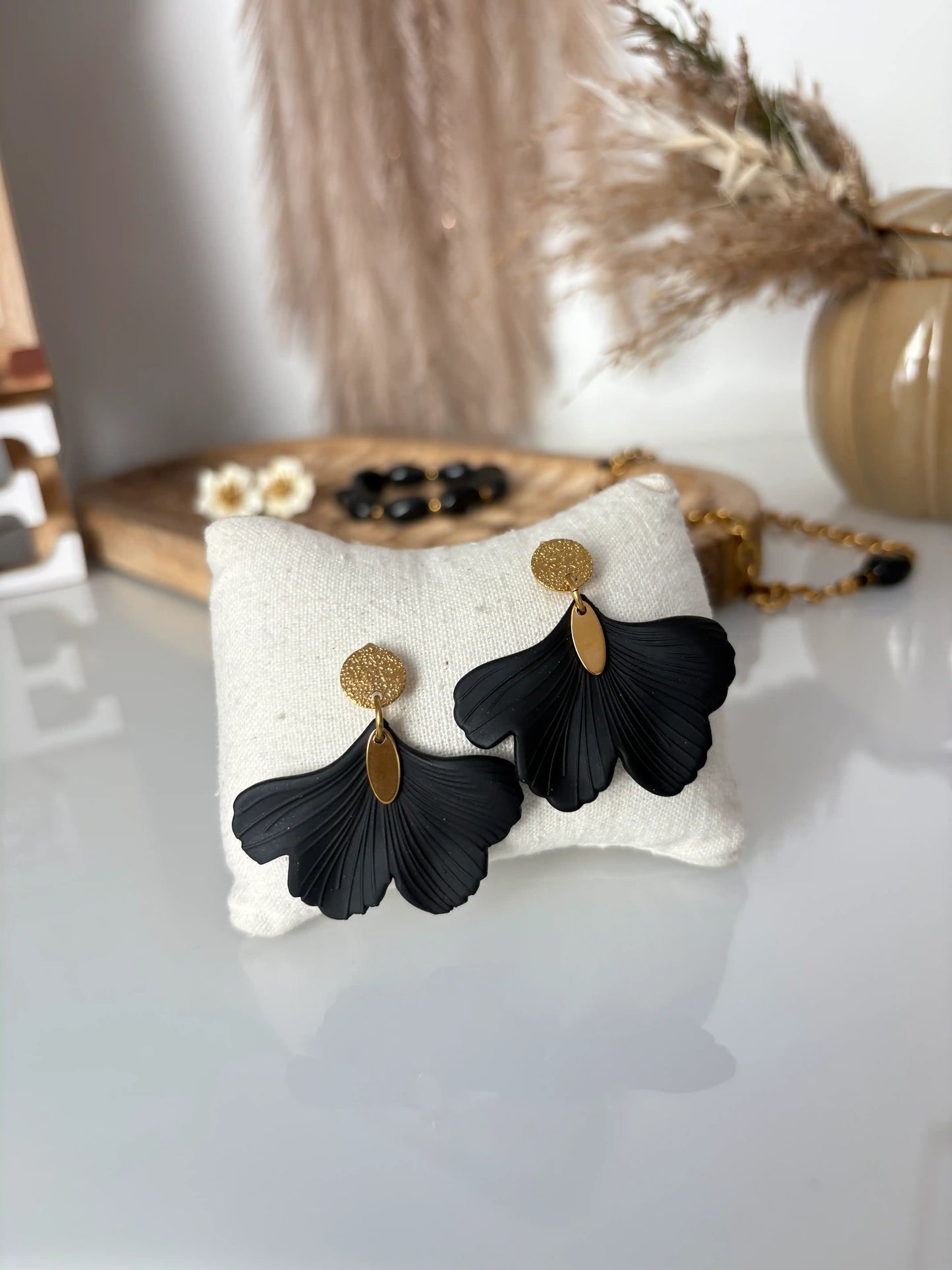 boucles d'oreilles à disque martelé décorées d'une feuille ginkgo noire surplombée d'un sequin ovale en acier inoxydable doré. Bijoux présentés sur un mini-coussin en coton beige.
