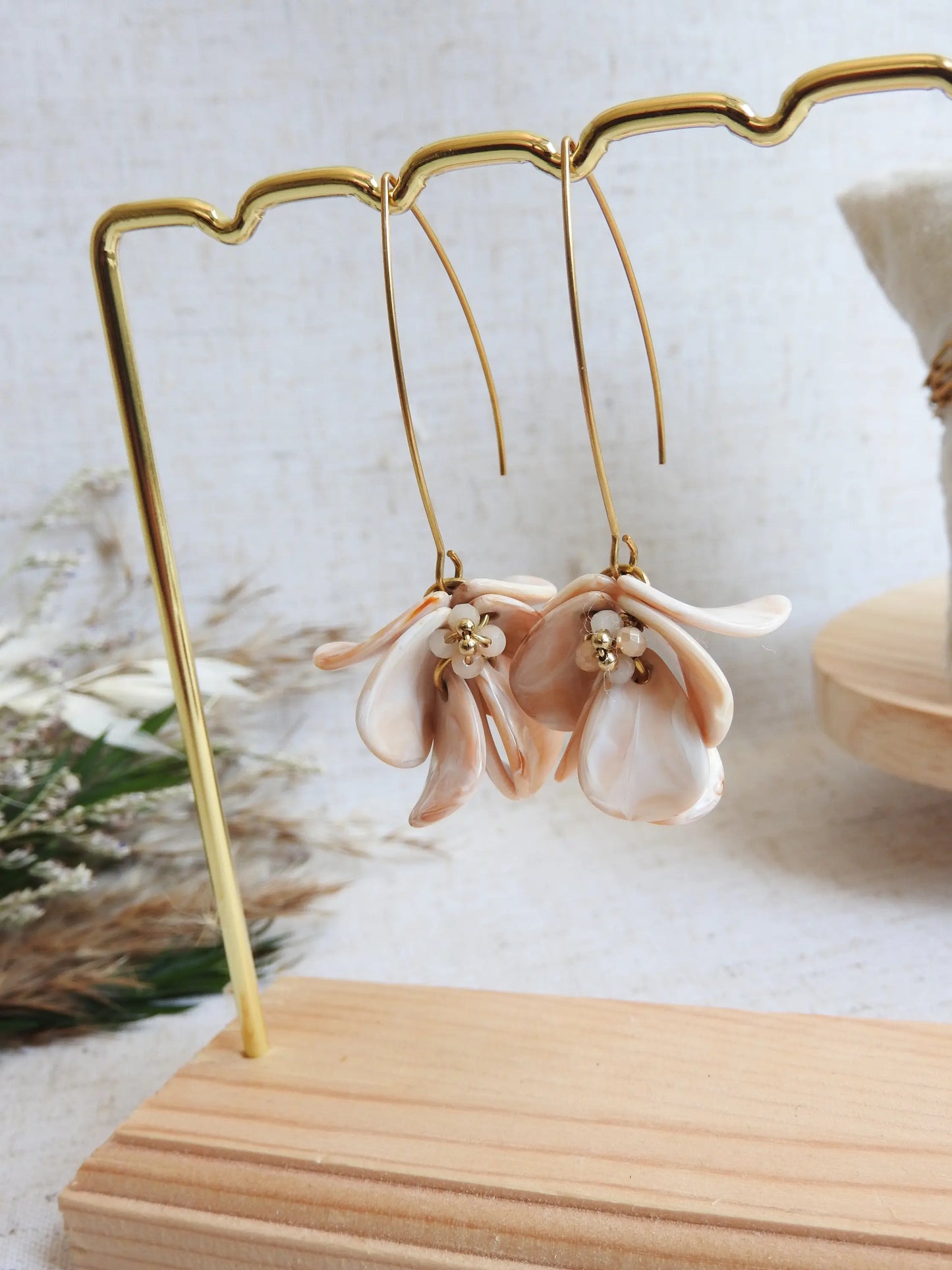 Boucles d'oreilles hameçon en acier inoxydable doré ornées de pétales beige à effet marbré, formant un bouquet floral. Bijoux agrémentés de perles de verre et acier doré, boucles suspendues sur un présentoir étallique doré et bambou posé sur une nappe beige décorée d'une brindille.