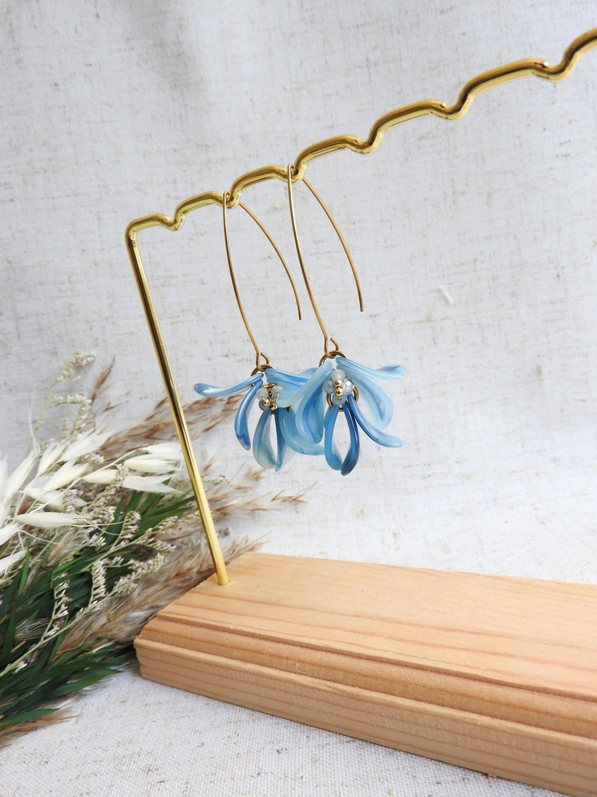 Boucles d'oreilles hameçon en acier inoxydable doré ornées de pétales bleu à effet marbré, formant un bouquet floral. Bijoux agrémentés de perles de verre et acier doré, boucles suspendues sur un présentoir métallique doré et bambou posé sur une nappe beige décorée d'une brindille.