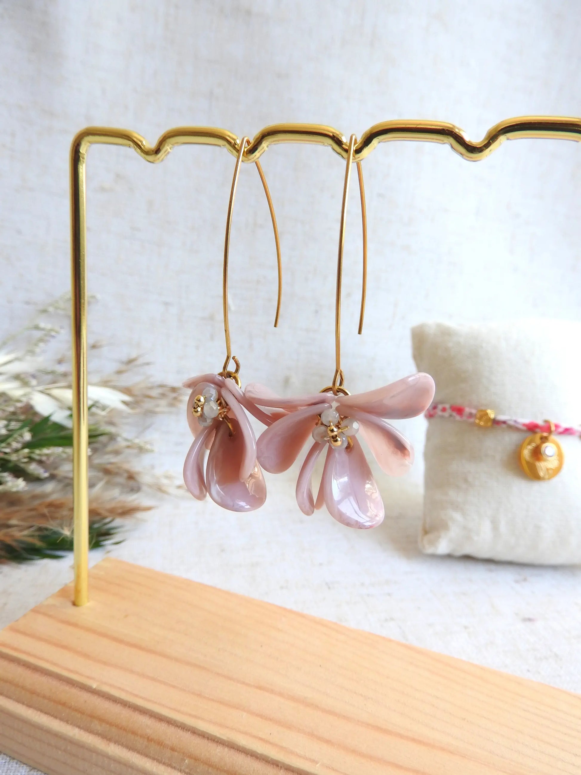 Boucles d'oreilles hameçon en acier inoxydable doré ornées de pétales rose à effet marbré, formant un bouquet floral. Bijoux agrémentés de perles de verre et acier doré, boucles suspendues sur un présentoir métallique doré et bambou posé sur une nappe beige décorée d'une brindille.