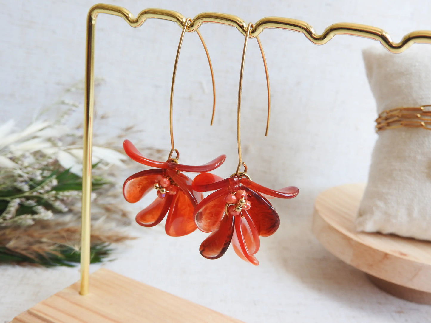 Boucles d'oreilles hameçon en acier inoxydable doré ornées de pétales rouge à effet marbré, formant un bouquet floral. Bijoux agrémentés de perles de verre et acier doré, boucles suspendues sur un présentoir métallique doré et bambou posé sur une nappe beige décorée d'une brindille.