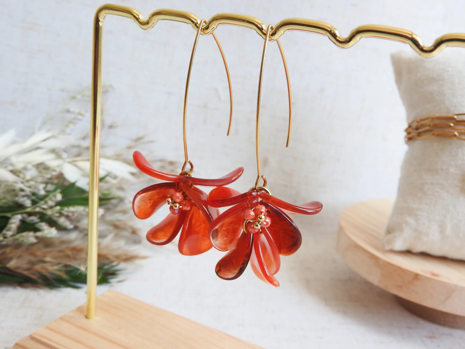 Boucles d'oreilles hameçon en acier inoxydable doré ornées de pétales rouge à effet marbré, formant un bouquet floral. Bijoux agrémentés de perles de verre et acier doré, boucles suspendues sur un présentoir métallique doré et bambou posé sur une nappe beige décorée d'une brindille.