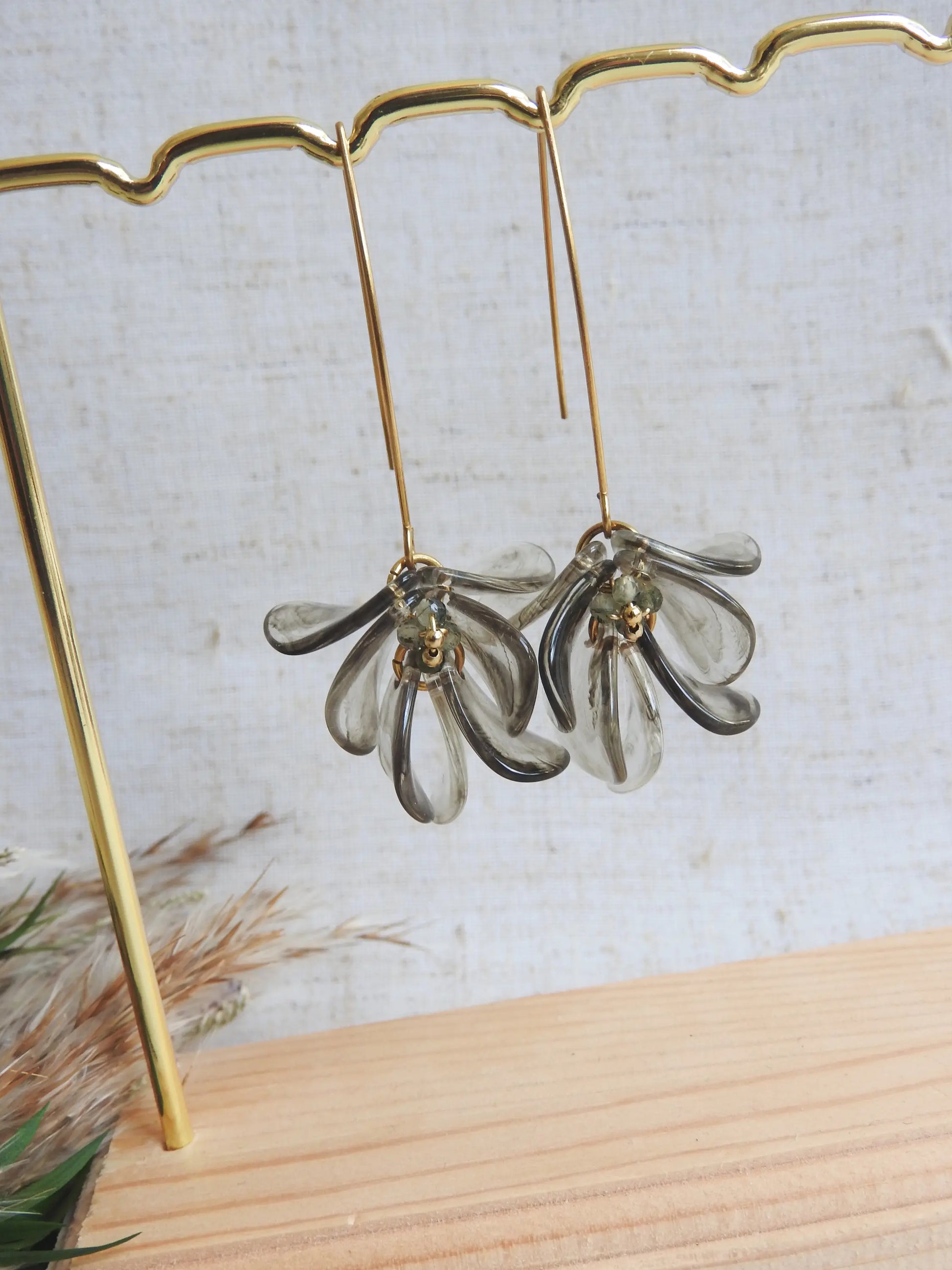 Boucles d'oreilles hameçon en acier inoxydable doré ornées de pétales vert kaki transparent formant un bouquet floral. Bijoux agrémentés de perles de verre et acier doré, boucles suspendues sur un présentoir métallique doré et bambou posé sur une nappe beige décorée d'une brindille.