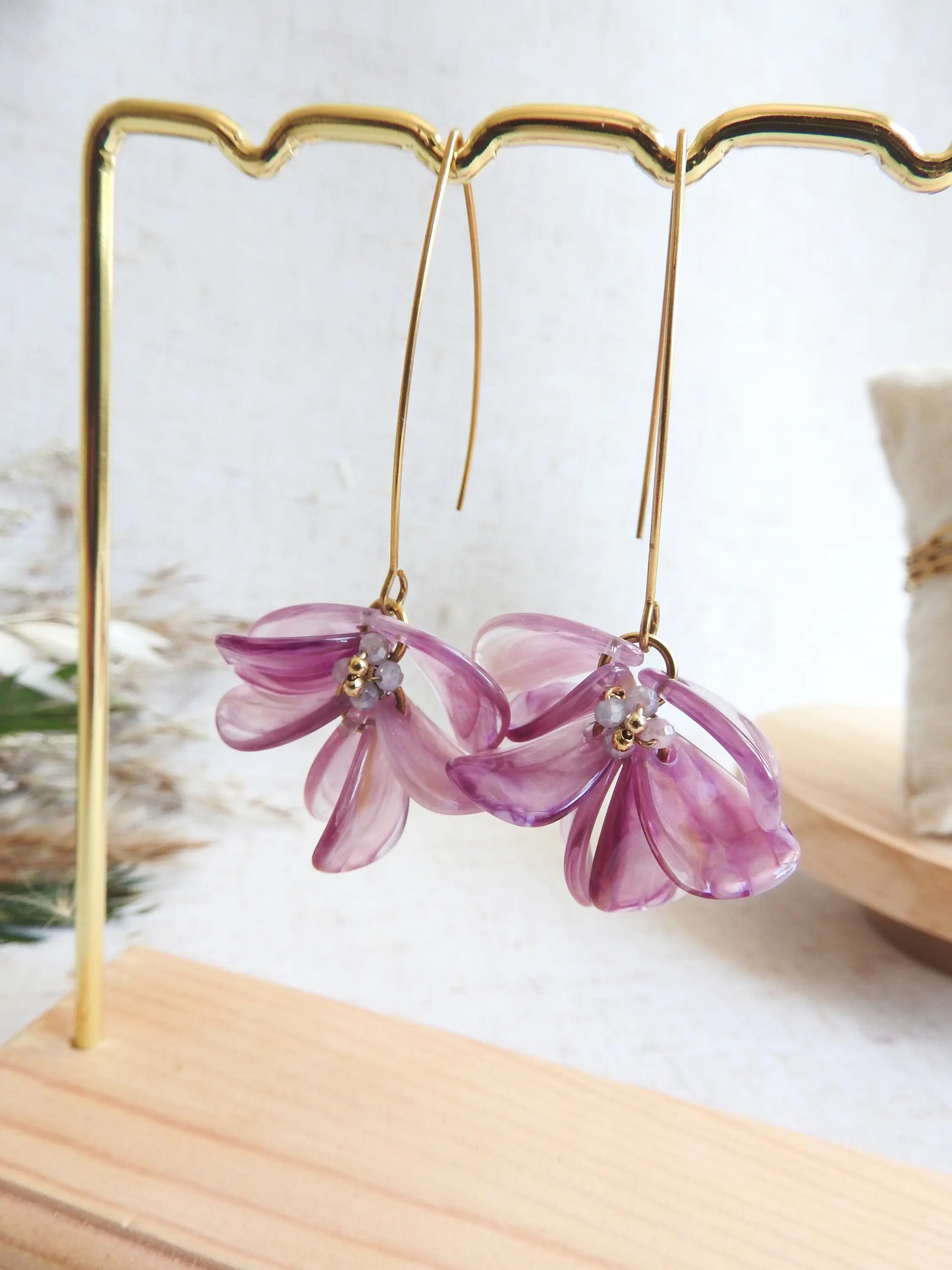 Boucles d'oreilles hameçon en acier inoxydable doré ornées de pétales violettes à effet marbré, formant un bouquet floral. Bijoux agrémentés de perles de verre et acier doré, boucles suspendues sur un présentoir métallique doré et bambou posé sur une nappe beige décorée d'une brindille.