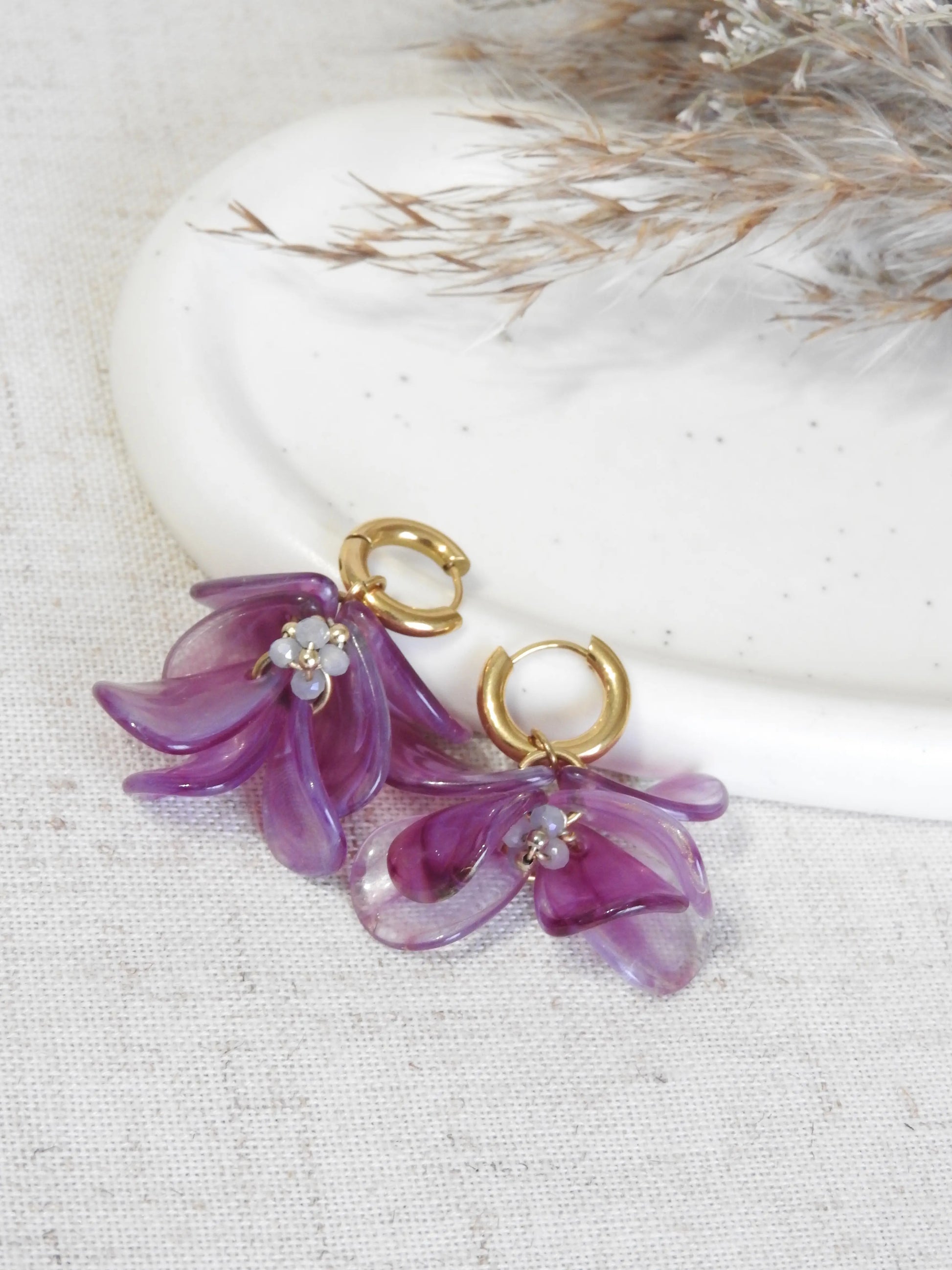 Boucles d'oreilles mini-créoles en acier inoxydable doré posé sur une nappe beige et une coupelle en céramique blanche. Bijou orné de pétales formant un bouquet floral en acrylique de couleur violette marbré.
