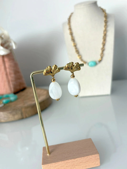 Boucles d'oreilles ornées d'une feuille de ginkgo en acier inoxydable doré avec une grande perle irrégulière blanche suspendue. Bijou présenté sur une support métallique doré et bambou posé sur une table avec en fond un buste présentoir présentant un collier.